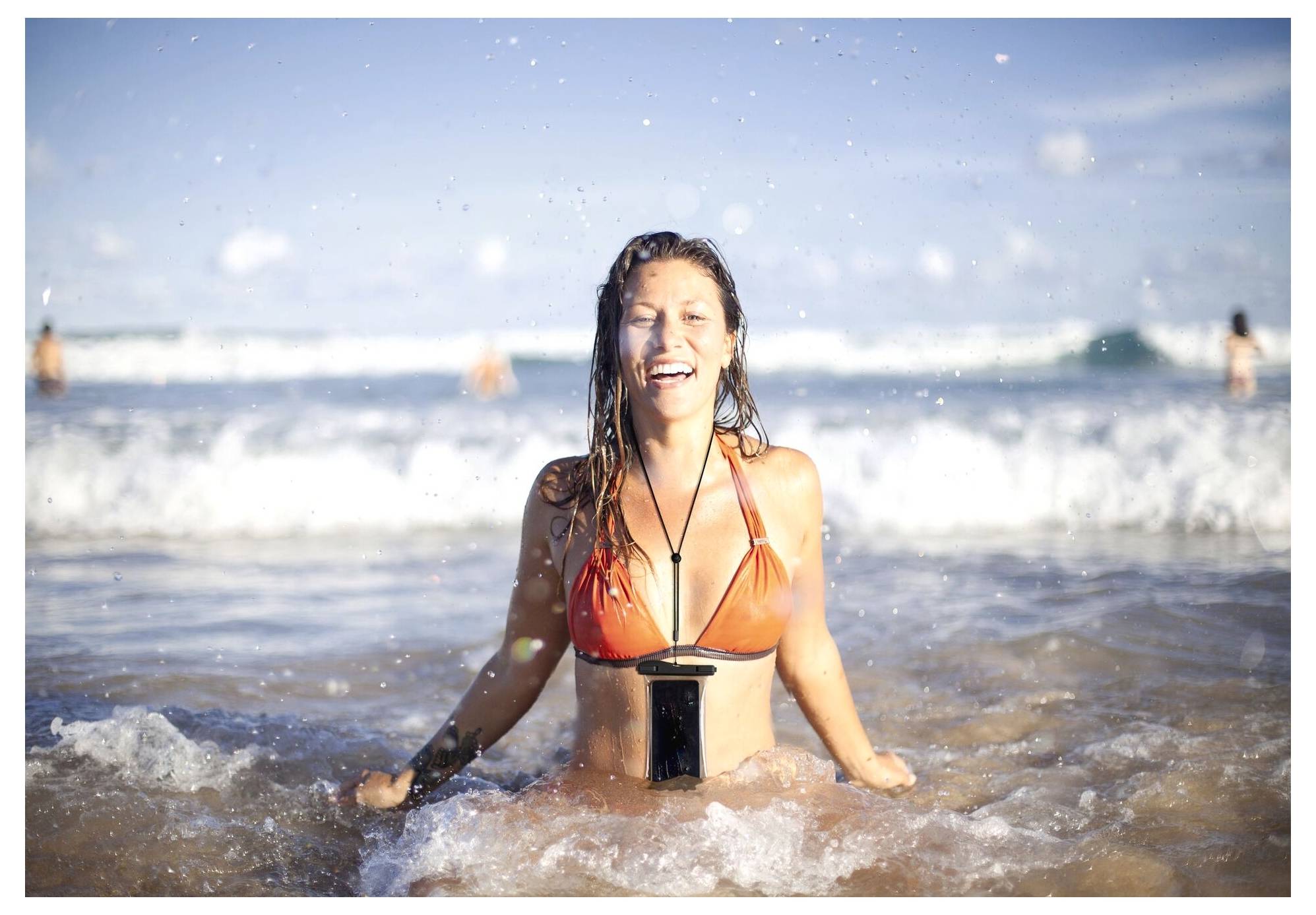 Eine lächelnde Person in einem orangefarbenen Bikini kniet in flachem Ozeanwasser mit Wellen und einem klaren blauen Himmel im Hintergrund.
