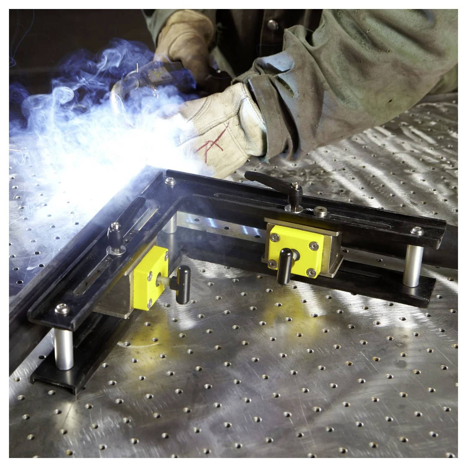 Eine Person, die Handschuhe und Schutzkleidung trägt, schweißt Metallteile auf einem gepunkteten Arbeitstisch, wobei helle Funken und Rauch sichtbar sind.