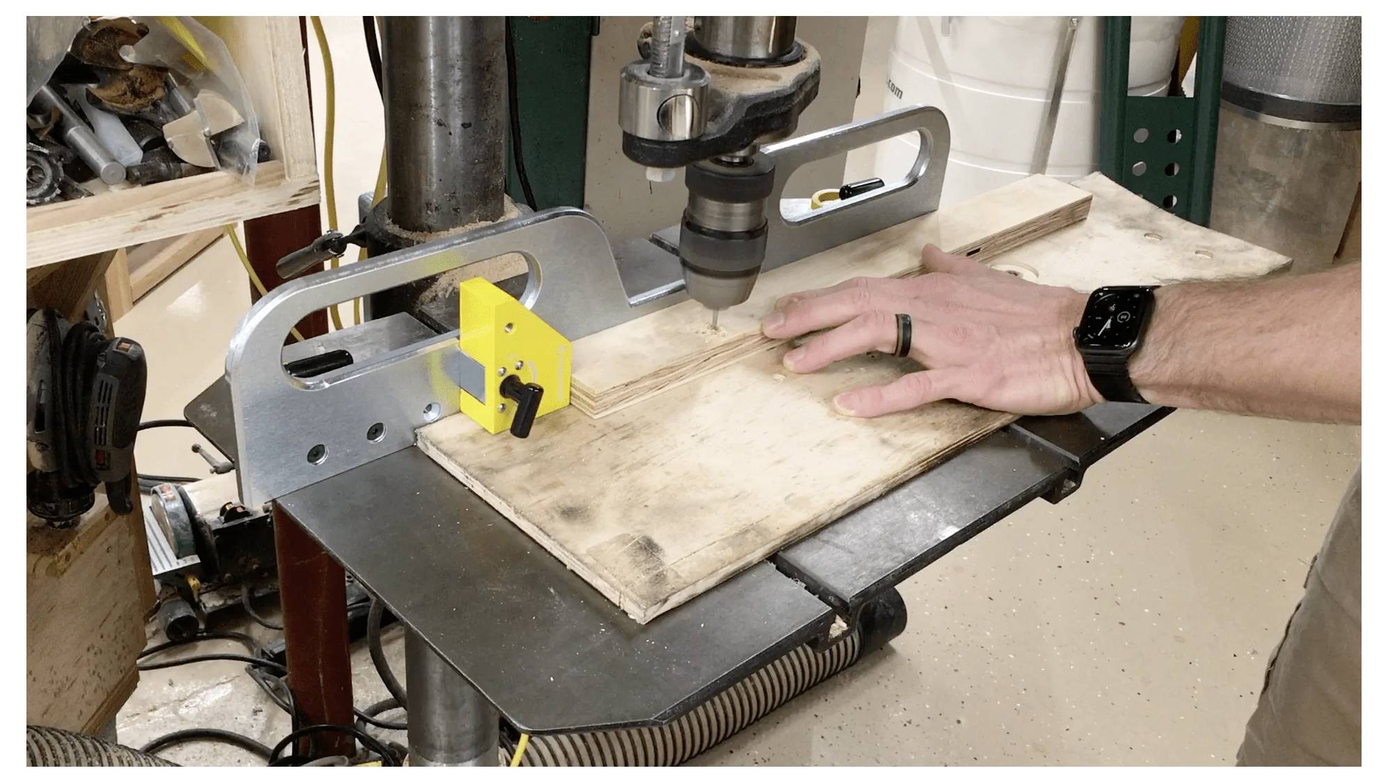 Eine Person, die an einer Bohrmaschine steht und mit einer Metallführung einen Schnitt in ein Holzstück macht und dieses dabei fest und sicher hält.