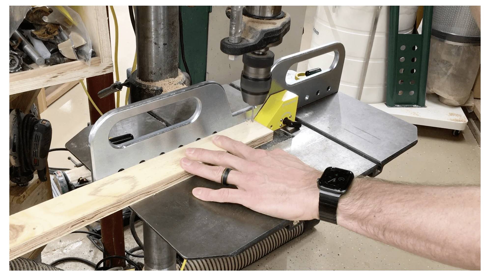 Eine Hand einer Person, die ein Holzbrett an einer Bohrmaschine in einer Werkstatt führt, trägt eine Smartwatch und einen Ring, mit Werkzeugen im Hintergrund.
