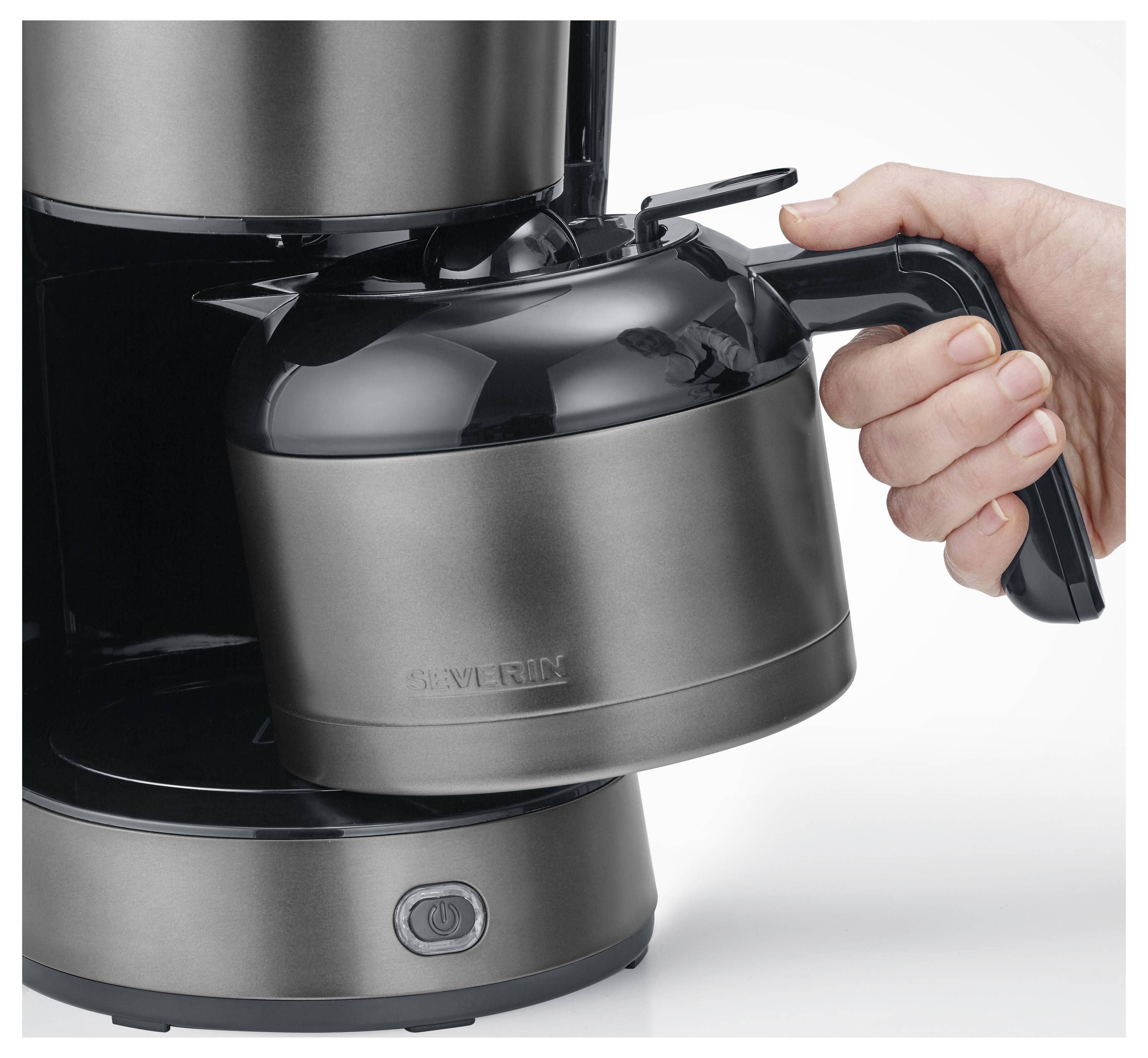 Eine Hand hält den Griff einer metallenen Kaffeekanne und stellt sie in einen Kaffeebereiter auf einer Küchenarbeitsplatte.