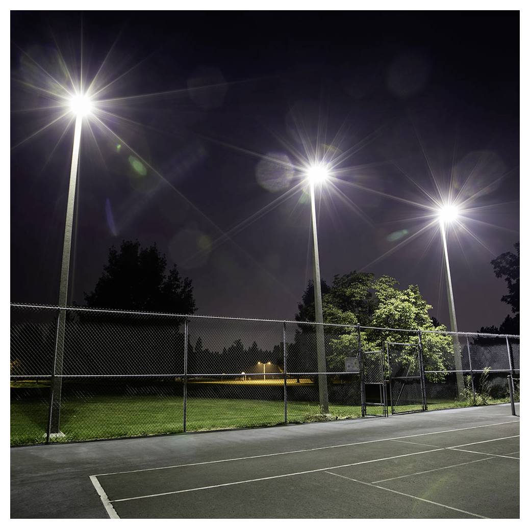 Ein Tennisplatz wird nachts von drei hohen Flutlichtern beleuchtet, die helles Licht über den umzäunten Bereich werfen. Im Hintergrund sind Bäume zu sehen.