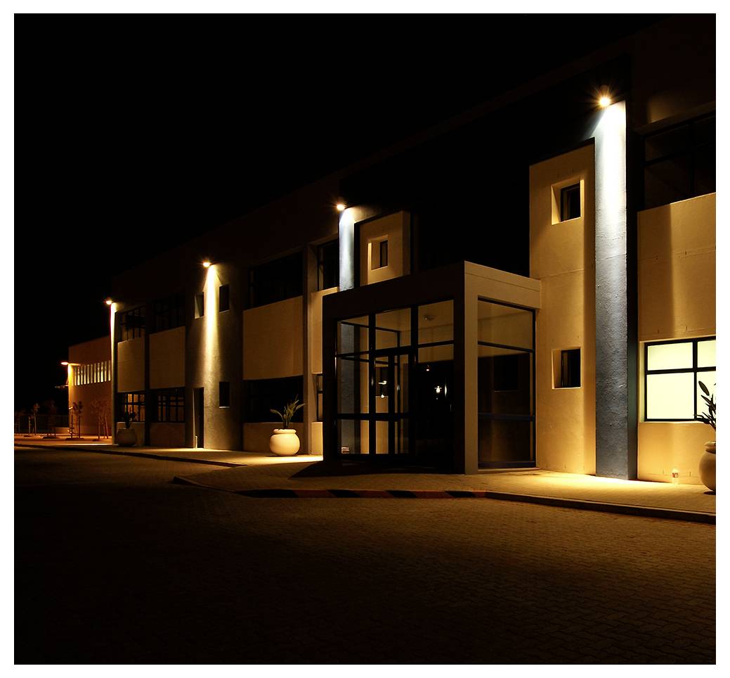 Eingang eines modernen Gebäudes bei Nacht, beleuchtet von Lichtern. Schatten und Highlights schaffen einen dramatischen Effekt an der Fassade.