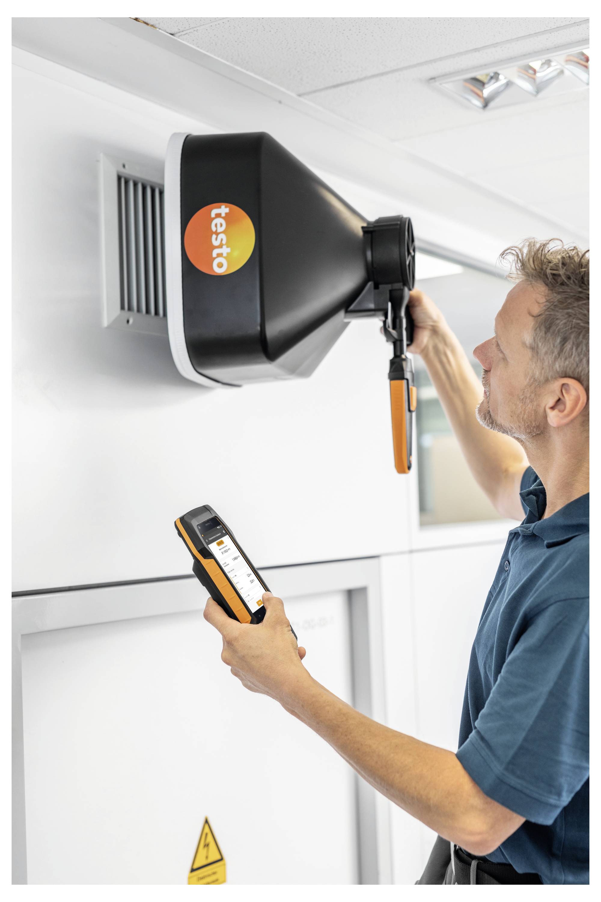 Eine Person benutzt ein tragbares Gerät, um den Luftstrom aus einem Belüftungskanal mit einem schwarzen Trichter-Werkzeug mit der Bezeichnung "testo" an einer Wand zu messen.