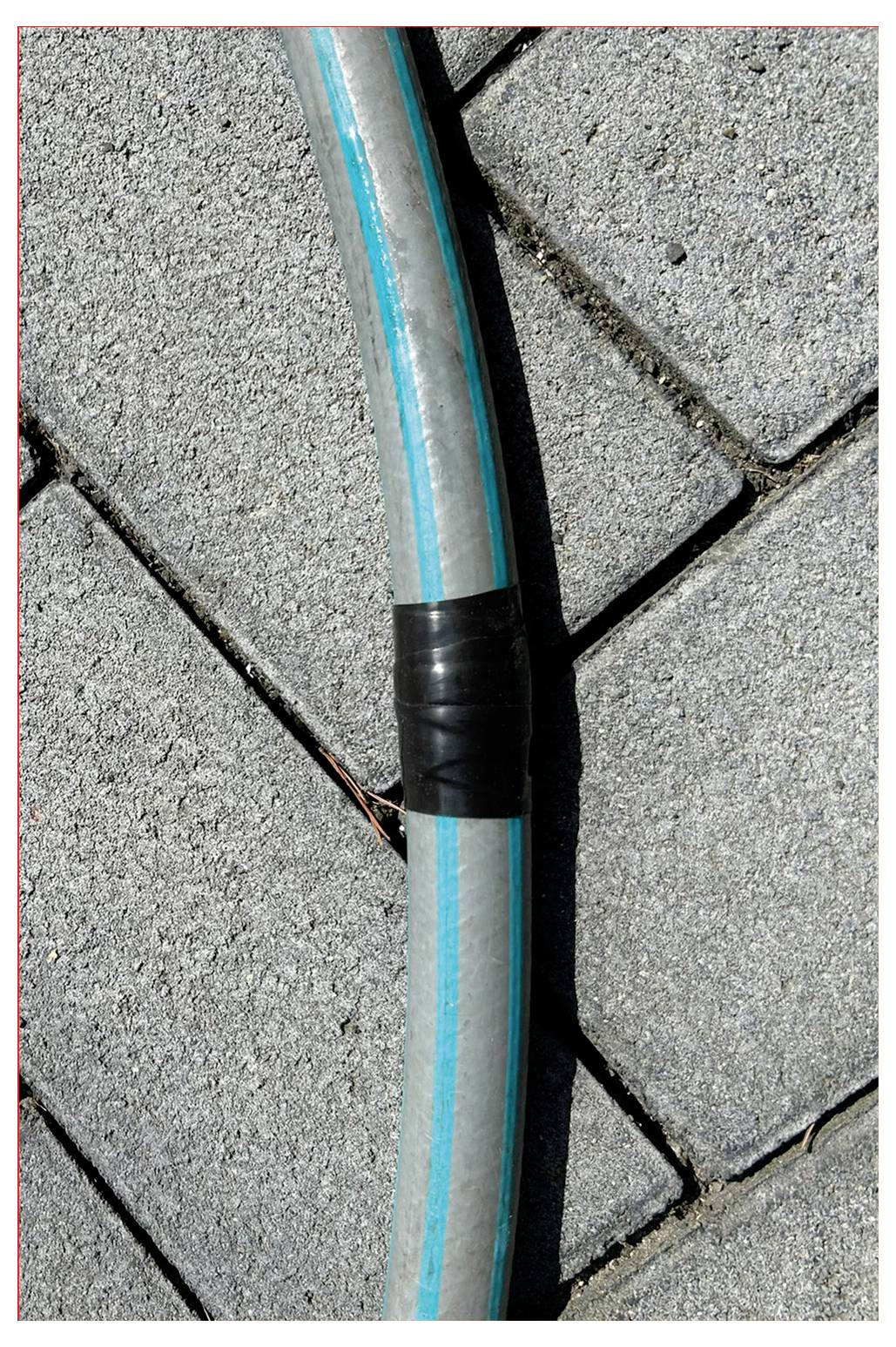 Ein grauer Schlauch mit einem blauen Streifen und schwarzem Klebeband führt über ein diagonal verlegtes graues Steinpflaster.
