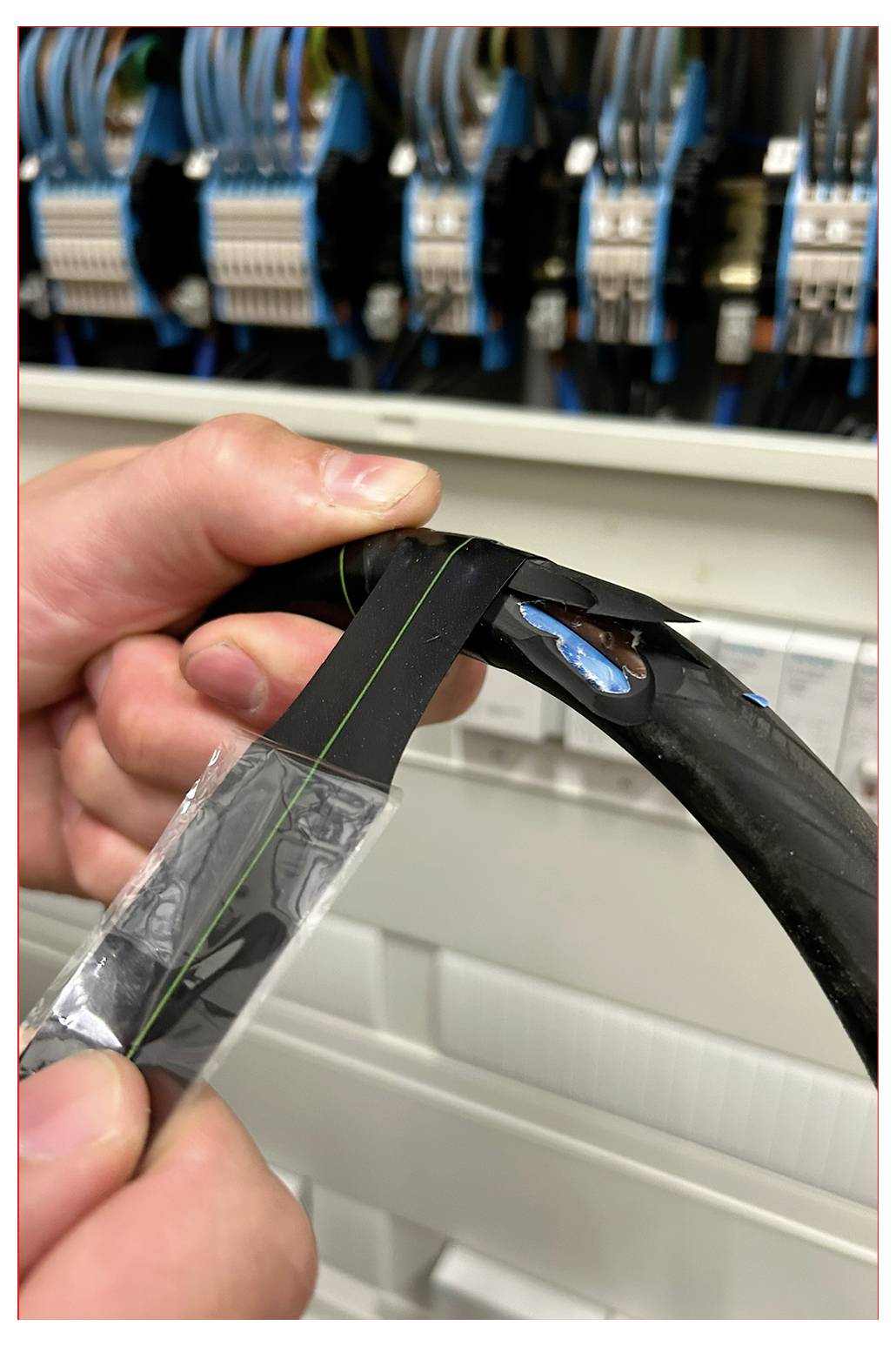 Nahaufnahme von Händen, die Isolierband um ein Kabel wickeln, vor einer Elektroverteilertafel mit Kabeln.