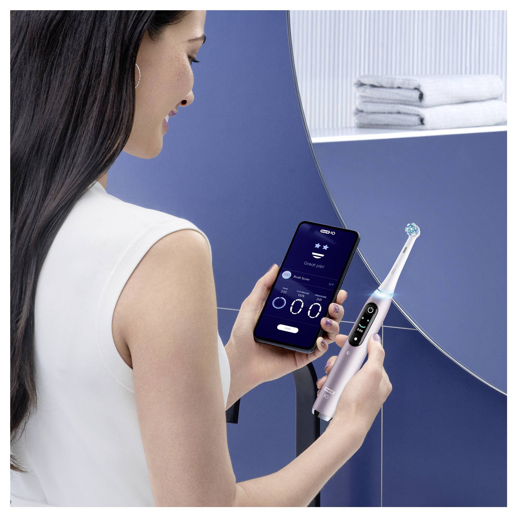 Eine Frau, die ein Smartphone hält, das mit einer elektrischen Zahnbürste gekoppelt ist, und App-Funktionen wie Reinigungsmodi zeigt. Ein Stapel Handtücher im Hintergrund.