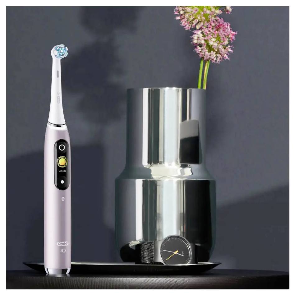 Eine elektrische Zahnbürste steht aufrecht neben einer silbernen Vase mit rosa Blumen und einer Armbanduhr, die auf einem schwarzen Tablett platziert ist.