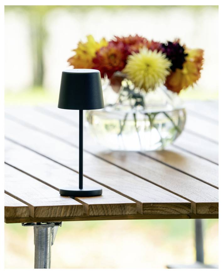 Eine schwarze Tischlampe auf einem Holztisch im Freien mit einer unscharfen Vase voller bunter Blumen im Hintergrund.