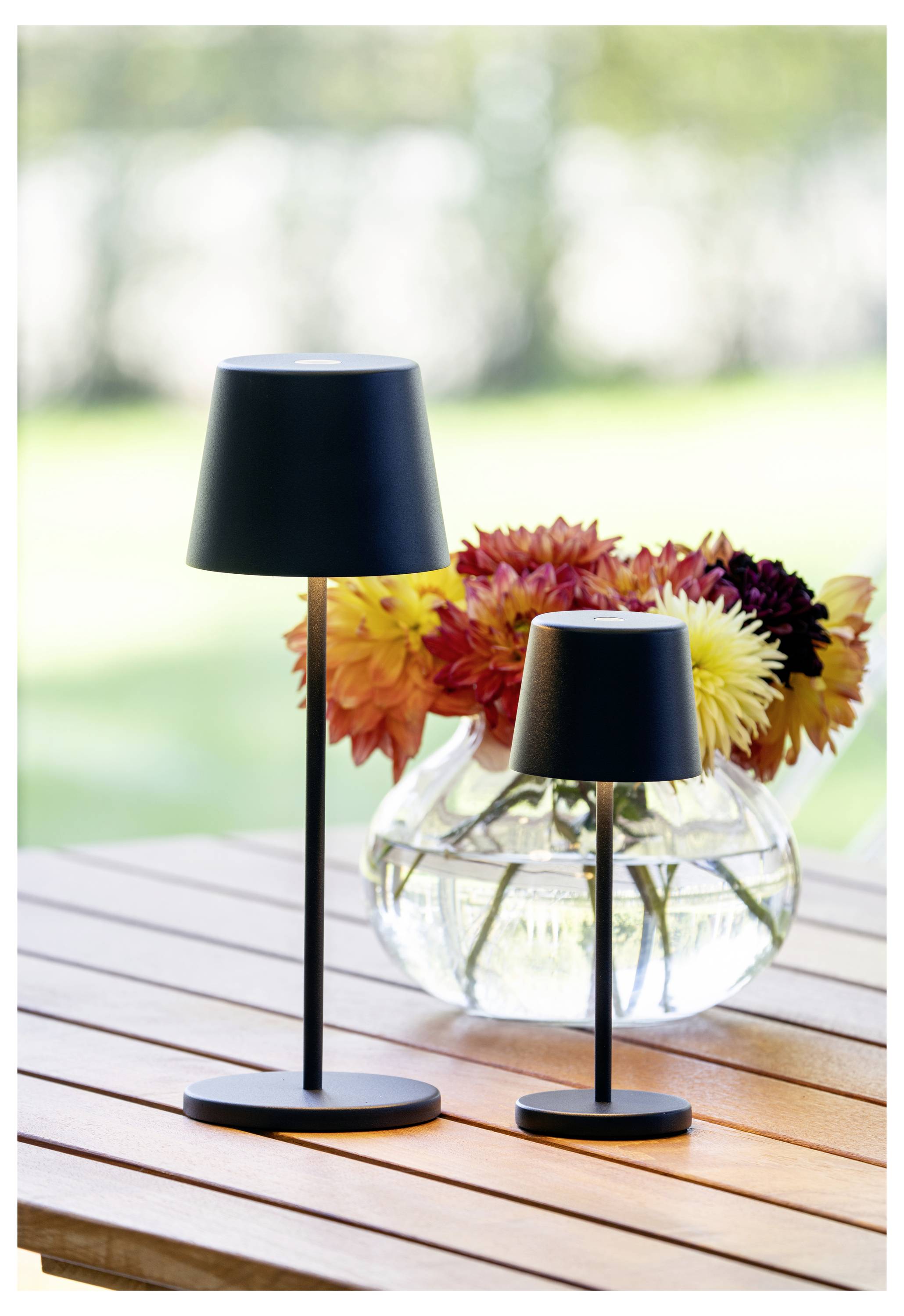Zwei schwarze Tischlampen unterschiedlicher Größe stehen auf einem Holztisch. Dahinter befindet sich eine klare Glasvase mit bunten Blumen.