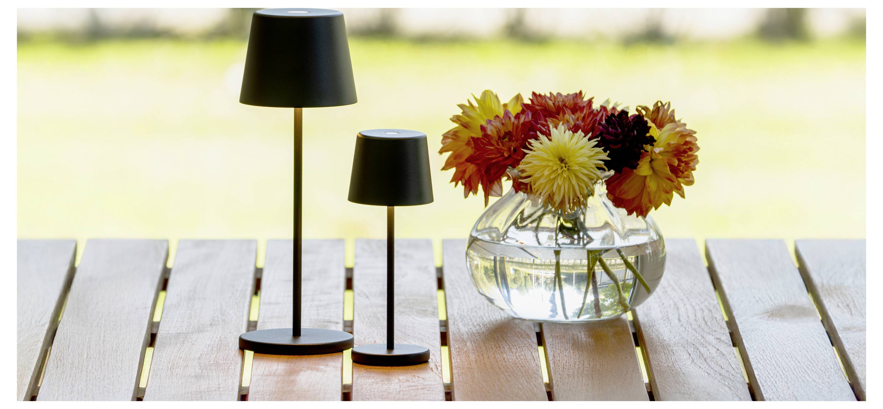 Ein Holztisch trägt eine Glasvase mit bunten Blumen und zwei kleinen, schwarzen stehenden Lampen vor einem unscharfen Hintergrund im Freien.