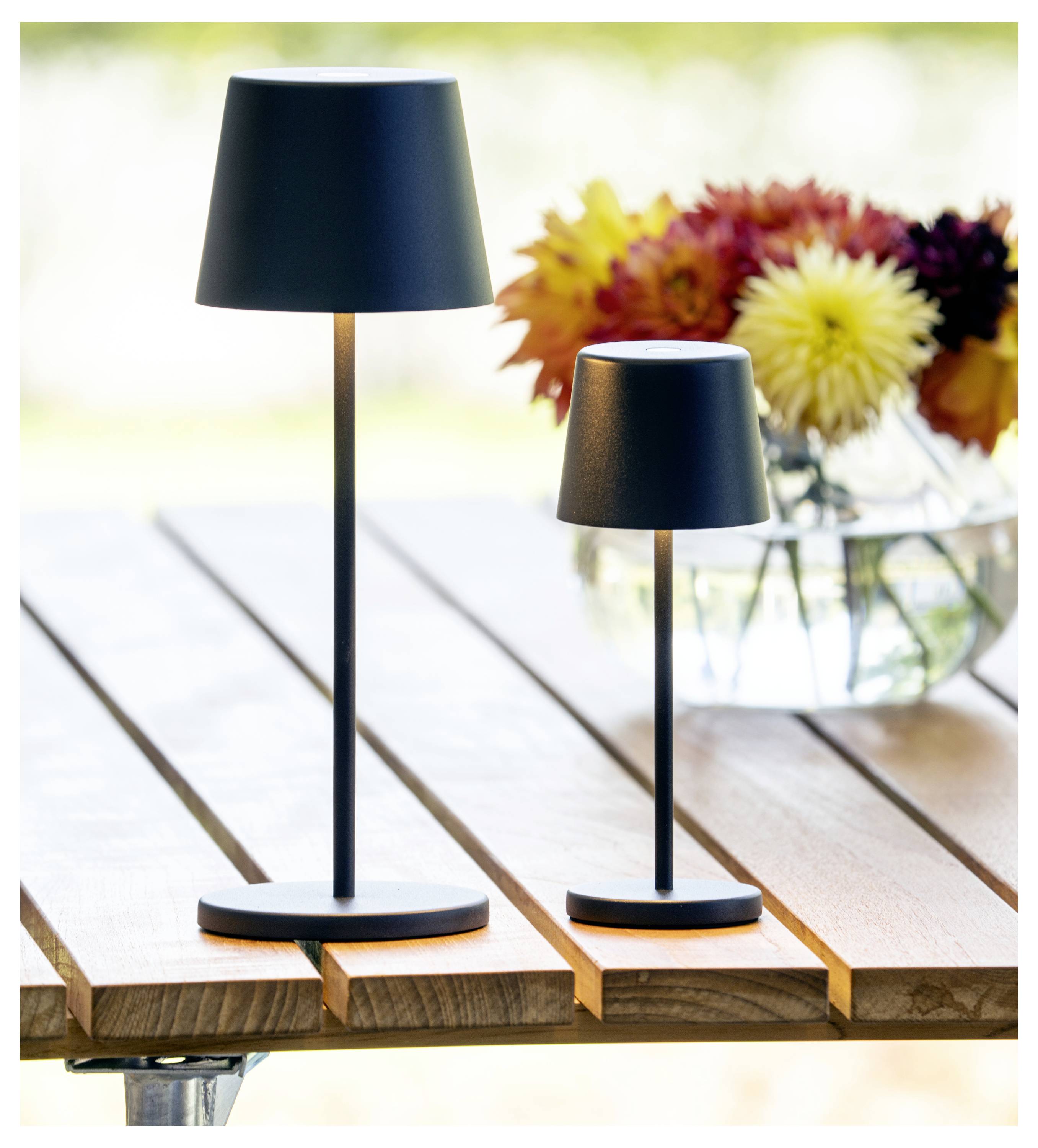 Zwei schwarze Lampen unterschiedlicher Größe auf einem Holztisch, mit einem unscharfen Hintergrund und einer Vase mit bunten Blumen in der Ferne.