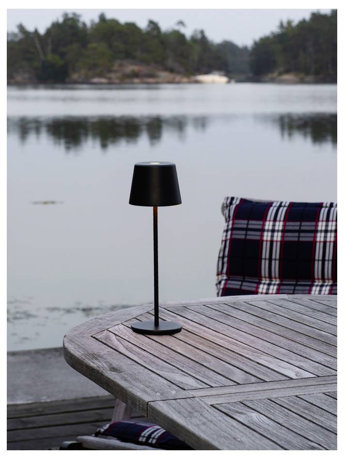 Eine kleine schwarze Lampe auf einem Holztisch im Freien, mit einem See und einer bewaldeten Uferline im Hintergrund. Eine karierte Decke ist über einen Stuhl gelegt.