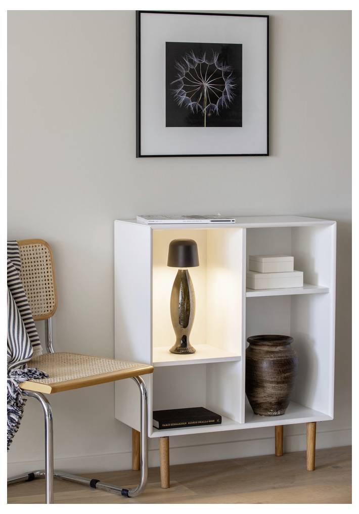 Ein kleiner weißer Schrank mit einer dekorativen Lampe und einer Vase, neben einem Stuhl. Darüber hängt ein gerahmtes Foto einer Pusteblume an der Wand.