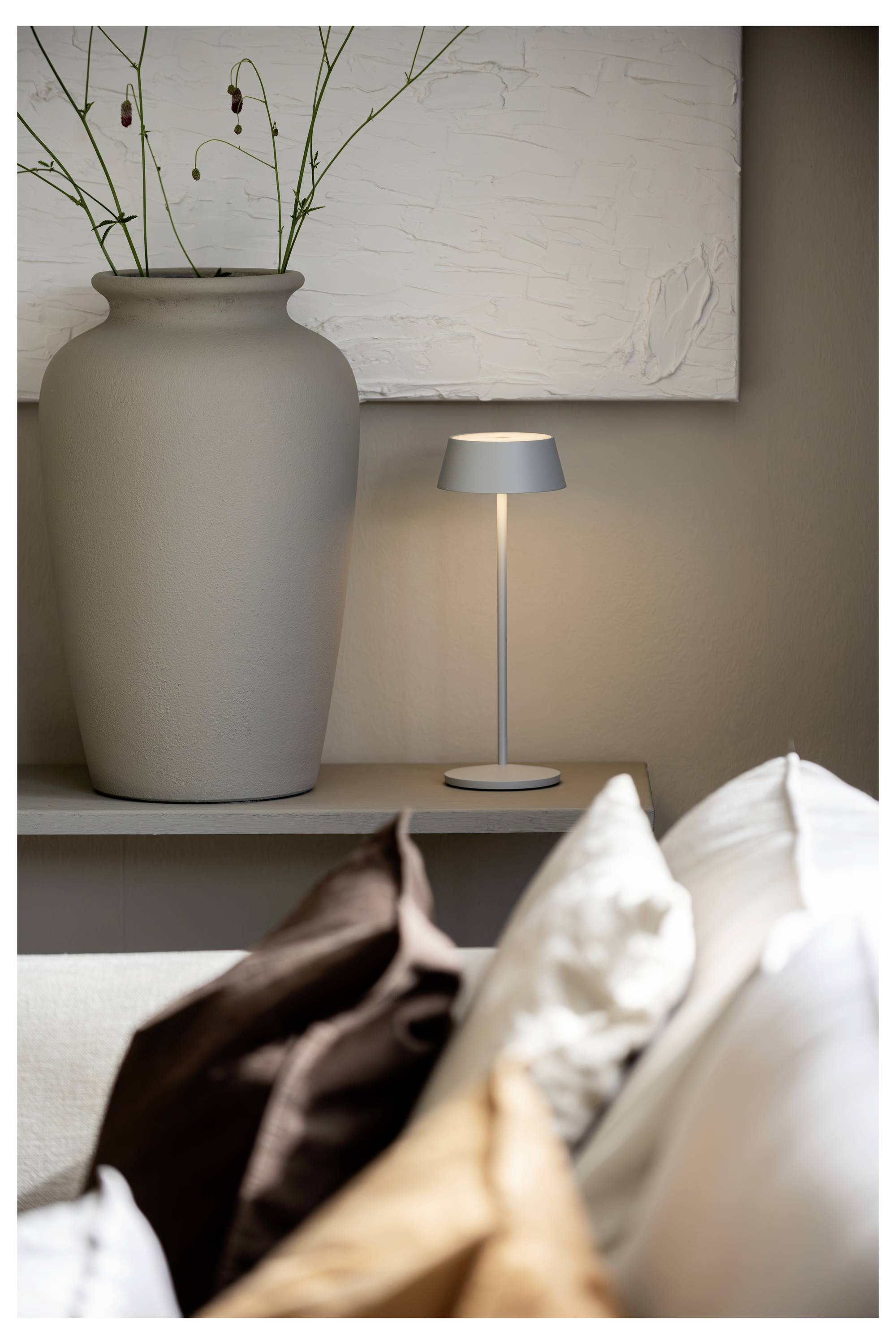 Ein modernes Wohnzimmer mit einer großen grauen Vase neben einer beleuchteten Tischlampe auf einem Regal und einem Sofa mit braunen und weißen Kissen im Vordergrund.