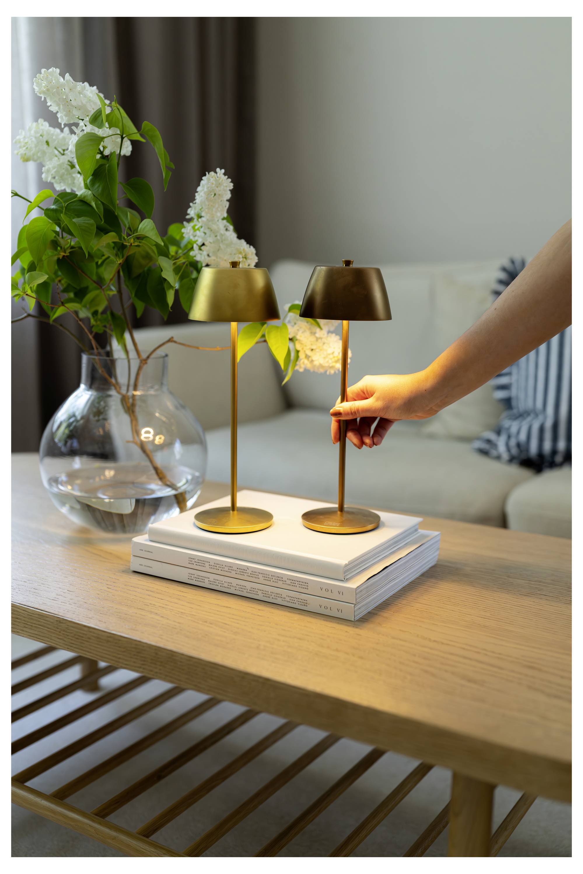 Eine Hand justiert eine kleine Messinglampe auf einem Holztisch neben einer größeren passenden Lampe und einer Vase mit weißen Blumen. Couch im Hintergrund.