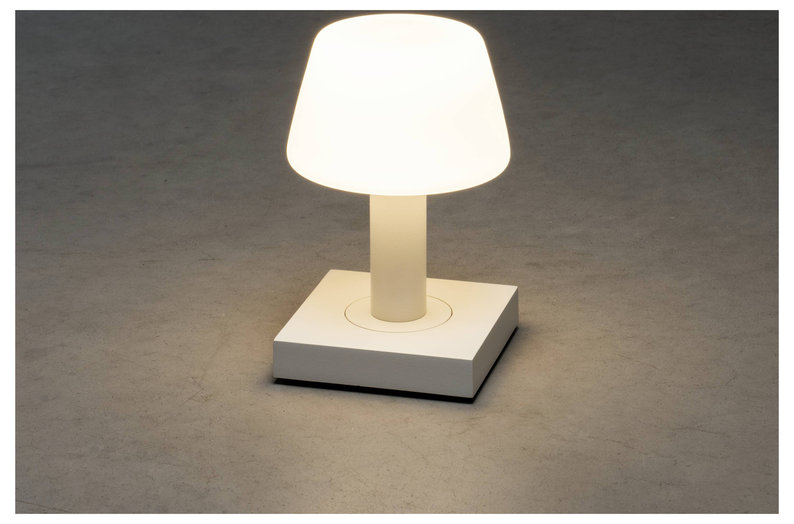 Eine kleine, moderne Tischlampe mit einem weißen Lampenschirm und einer quadratischen Basis strahlt ein sanftes, warmes Licht in einem dämmrig beleuchteten Raum.