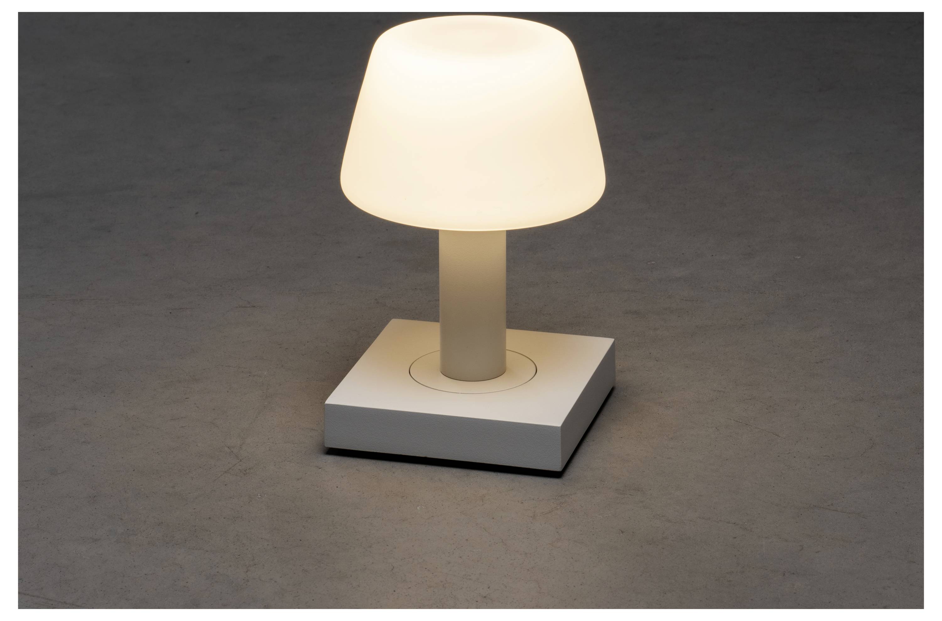 Eine kleine, moderne Tischlampe mit einem weißen Lampenschirm verbreitet ein warmes Leuchten, die auf einer grauen Oberfläche steht und eine minimalistische und gemütliche Atmosphäre schafft.