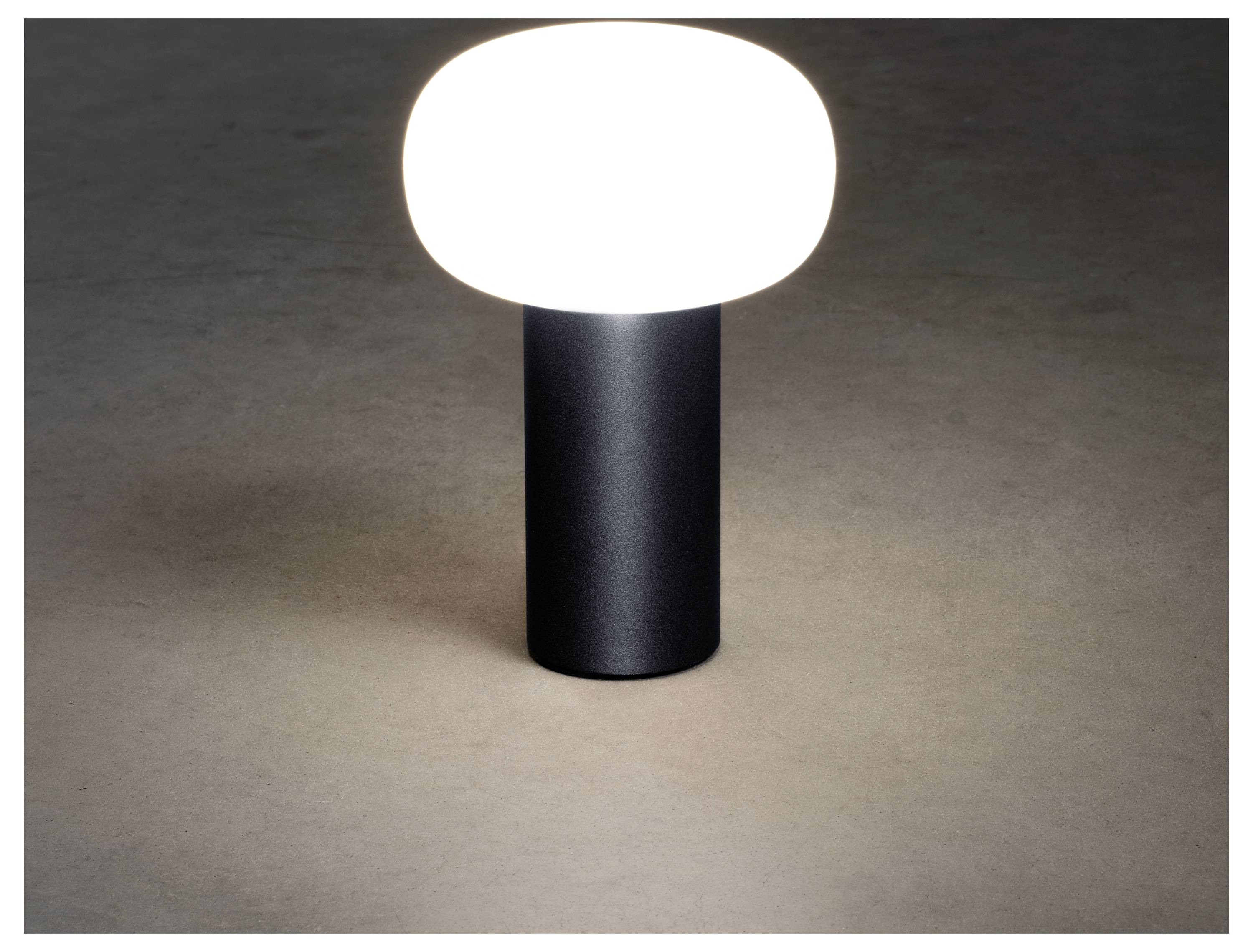 Eine moderne Tischlampe mit einem runden, leuchtend weißen Schirm auf einem zylindrischen schwarzen Fuß beleuchtet eine neutrale Oberfläche.