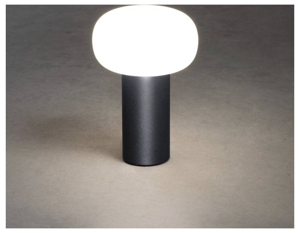 Eine moderne Tischlampe mit einer abgerundeten, weißen Oberseite verbreitet ein warmes Leuchten und steht auf einer glatten, dunklen zylindrischen Basis vor einem neutralen Hintergrund.