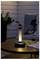 Eine moderne Tischlampe strahlt warmes Licht auf einen Gartentisch, neben zwei leeren Weingläsern und einer Zeitschrift, während im Hintergrund eine Pflanze steht.