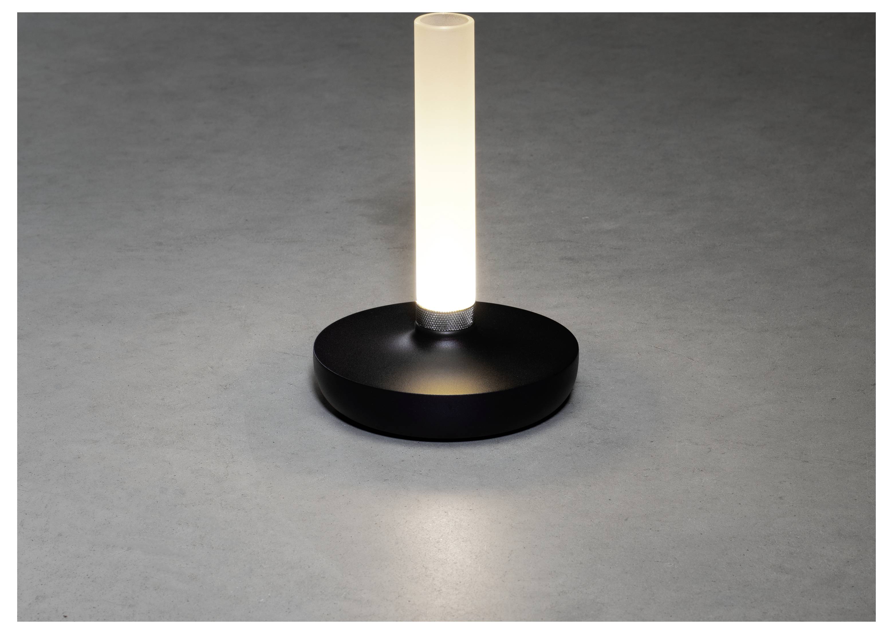 Eine kleine, moderne Tischlampe mit einem zylindrischen Leuchtenkörper auf einer runden schwarzen Basis, die ein weiches Licht auf die graue Oberfläche wirft.