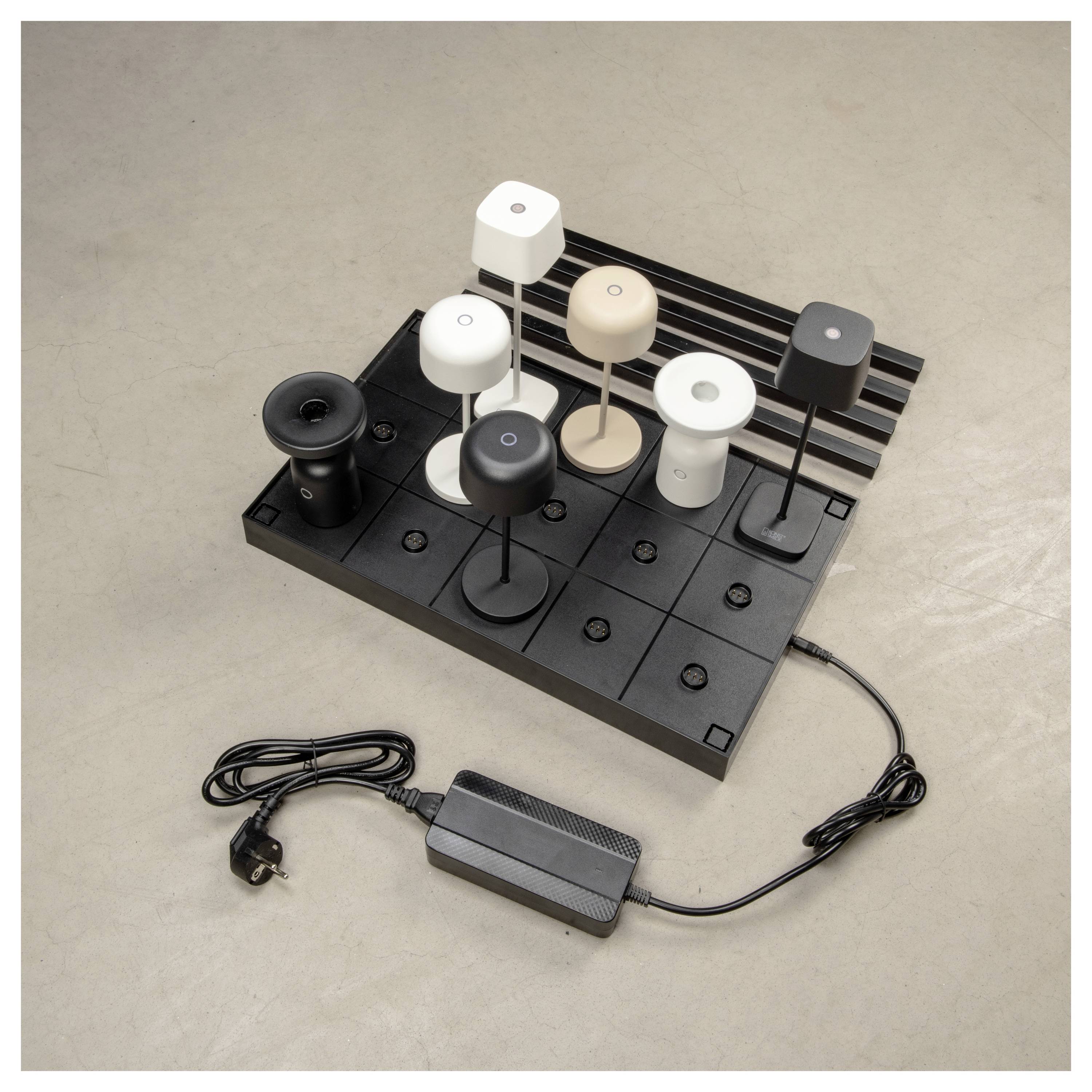 Eine schwarze Basis zeigt acht moderne Tischlampen in Weiß, Schwarz und Beige. Ein Netzteil mit Stecker ist mit der Basis verbunden.