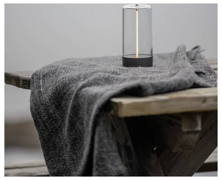 Eine kleine, zylindrische Lampe verbreitet ein weiches Licht und steht auf einer Holzbank, die mit einer grauen, strukturierten Decke bedeckt ist.