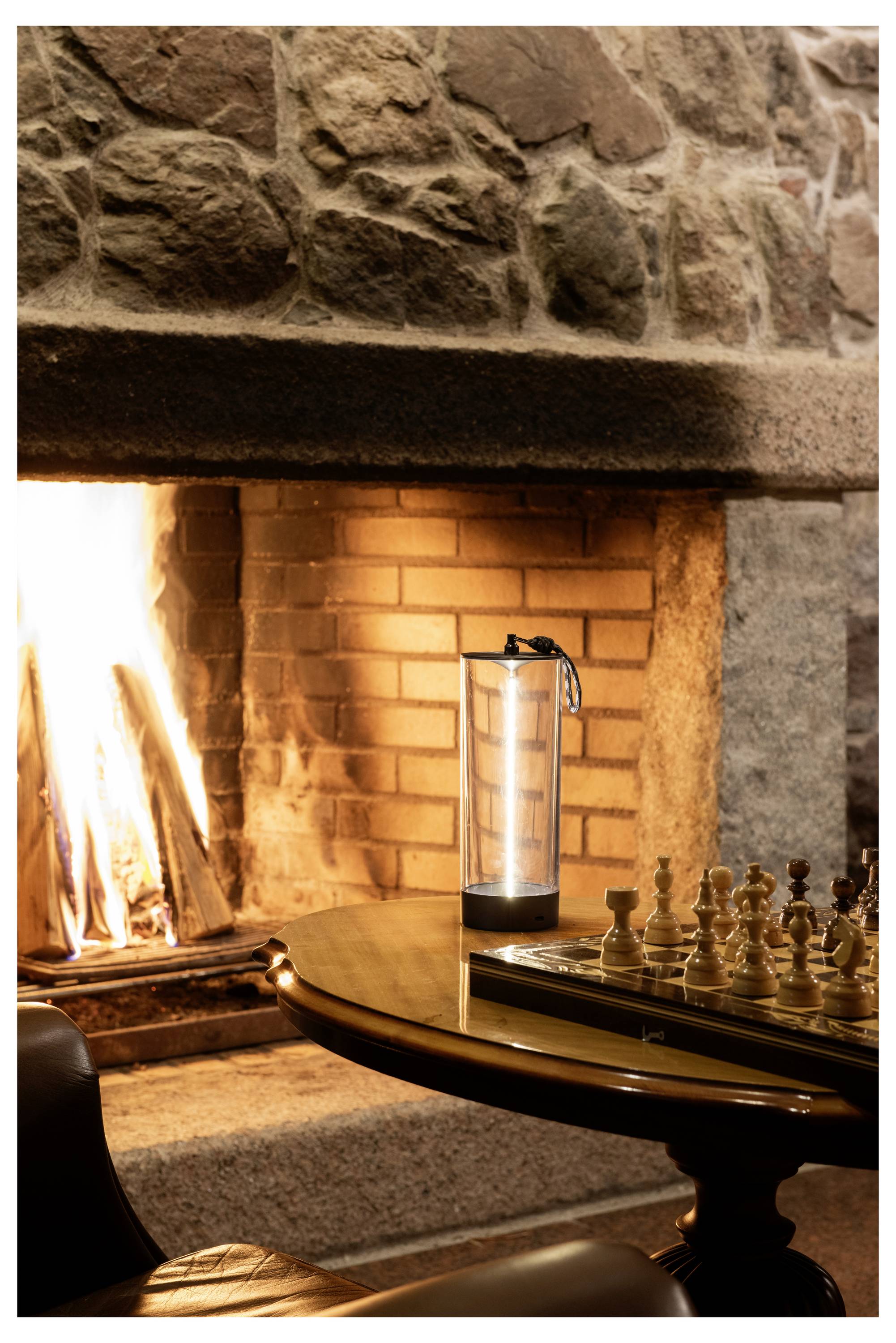 Eine gemütliche Wohnzimmerszene mit einem brennenden Kamin, einem hohen Wasserglas auf einem Holztisch und einem für das Spiel aufgebauten Schachbrett im Vordergrund.