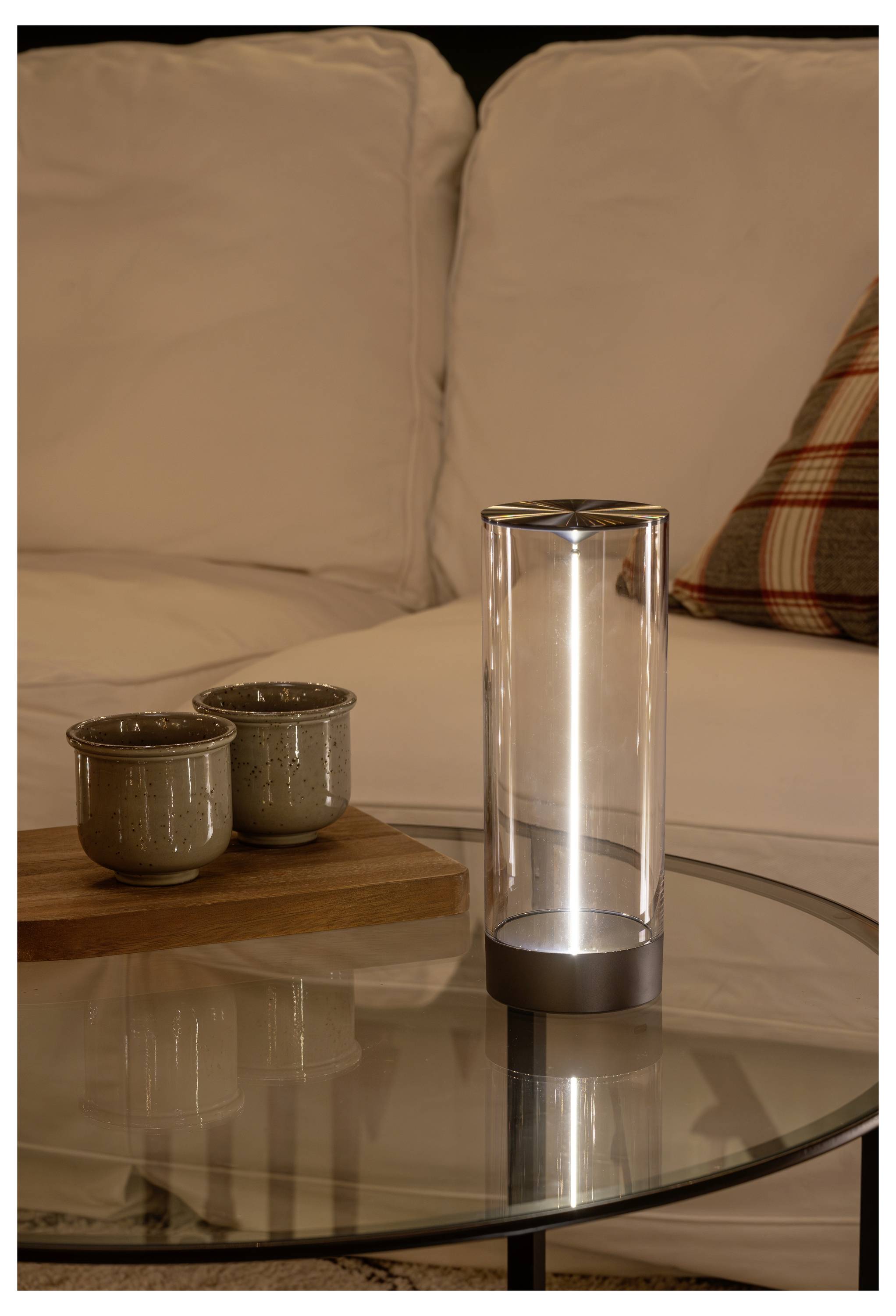 Eine zylindrische Glaslampe verbreitet ein weiches Licht auf einem runden Glastisch, mit zwei Tassen auf einem Holztablett, neben einem weißen Sofa mit einem karrierten Kissen.