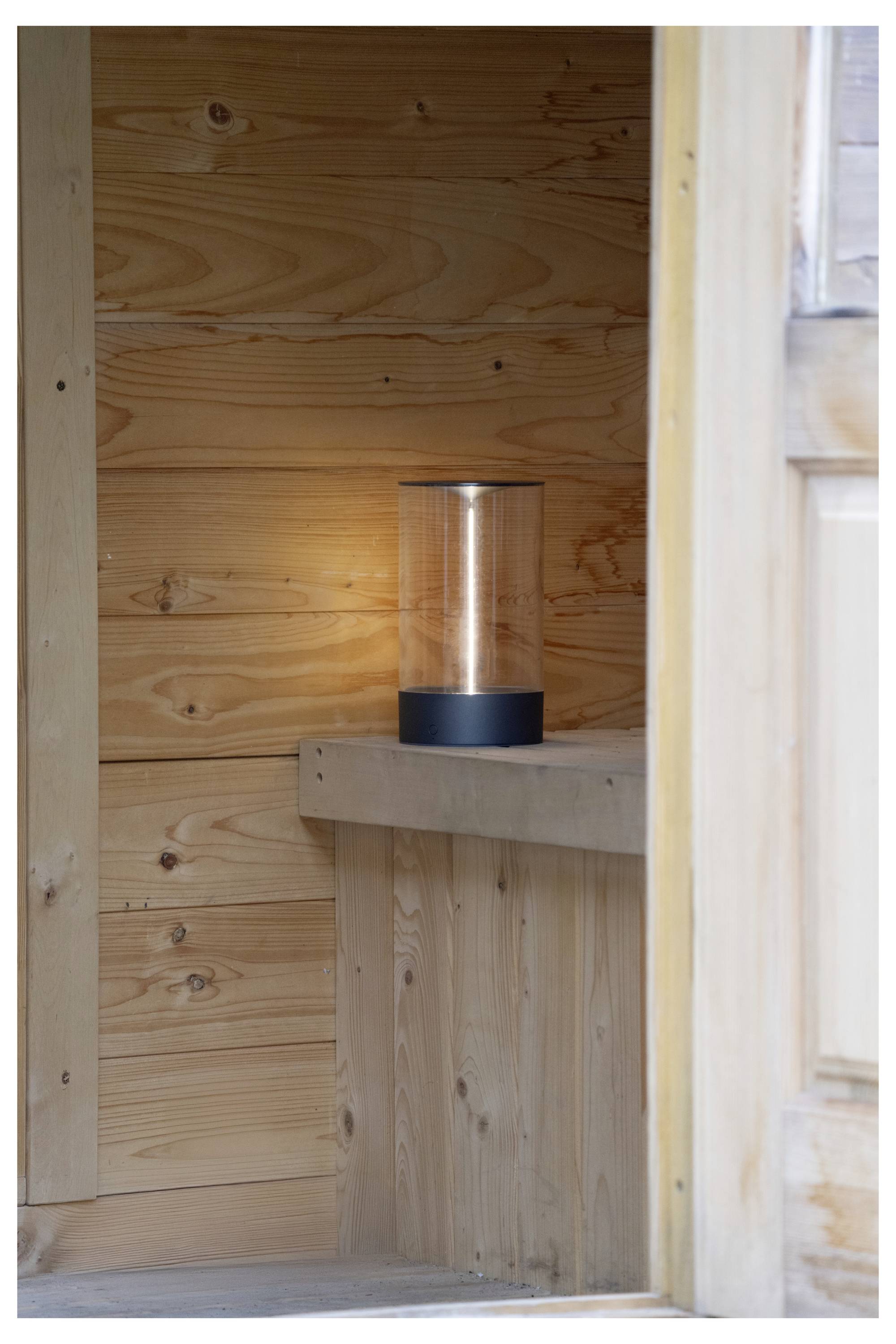 Eine zylindrische Laterne mit einem schwarzen Sockel verbreitet ein warmes Leuchten und steht auf einem Holzregal in einem mit Holzvertäfelung ausgestatteten Raum.