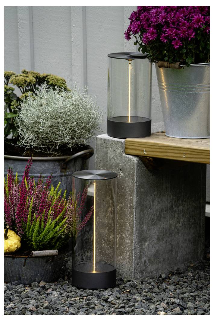 Outdoor-Szene mit zwei zylindrischen Laternen auf einem Kiesweg, umgeben von Topfblumen, einschließlich violetter Blüten in einem Metalleimer.