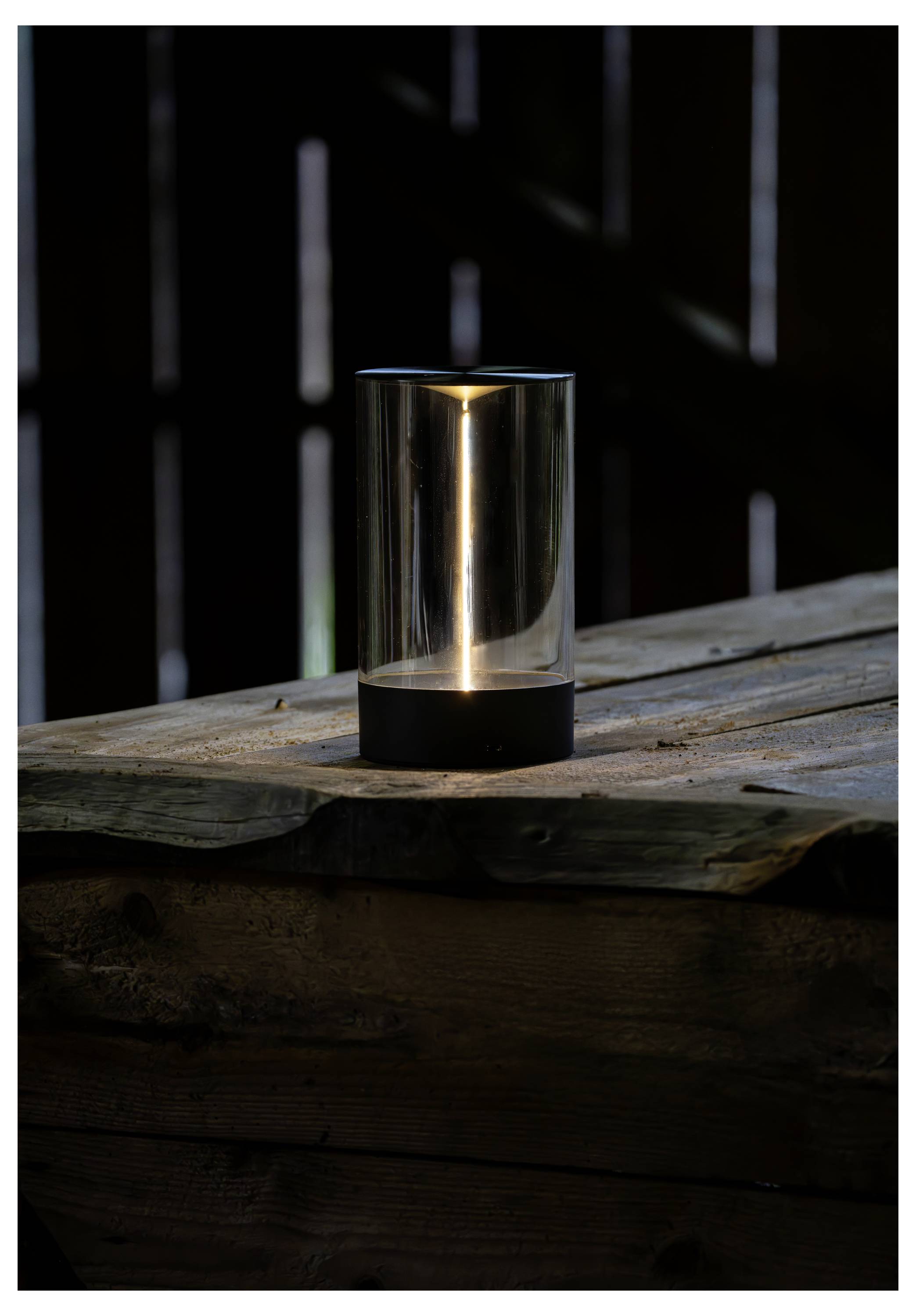 Eine kleine, zylindrische Lampe mit schwarzem Sockel und transparentem Deckel steht auf einer rustikalen Holzoberfläche und verbreitet ein warmes, weiches Licht.