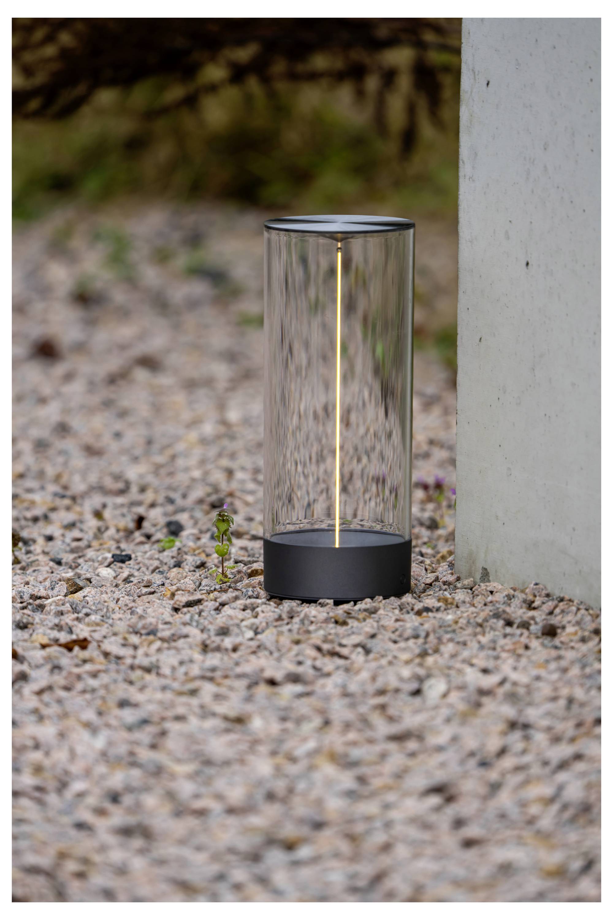 Eine moderne, zylindrische Außenlampe mit transparentem Gehäuse, die ein weiches Licht abstrahlt, steht auf einer Kiesfläche neben einem Betonpfosten.