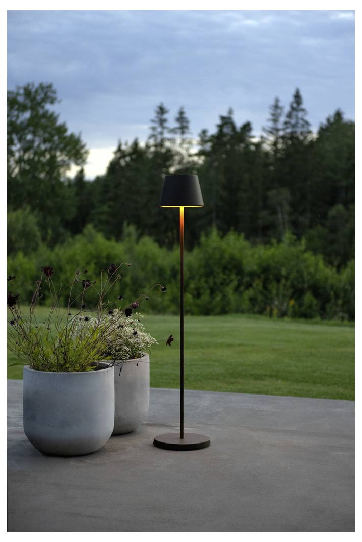Eine elegante Außenlampe leuchtet auf einer Terrasse, vor einer Kulisse aus üppig grünen Bäumen und einem bewölkten Himmel in der Abenddämmerung. Zwei graue Pflanzgefäße mit Blattwerk befinden sich in der Nähe.