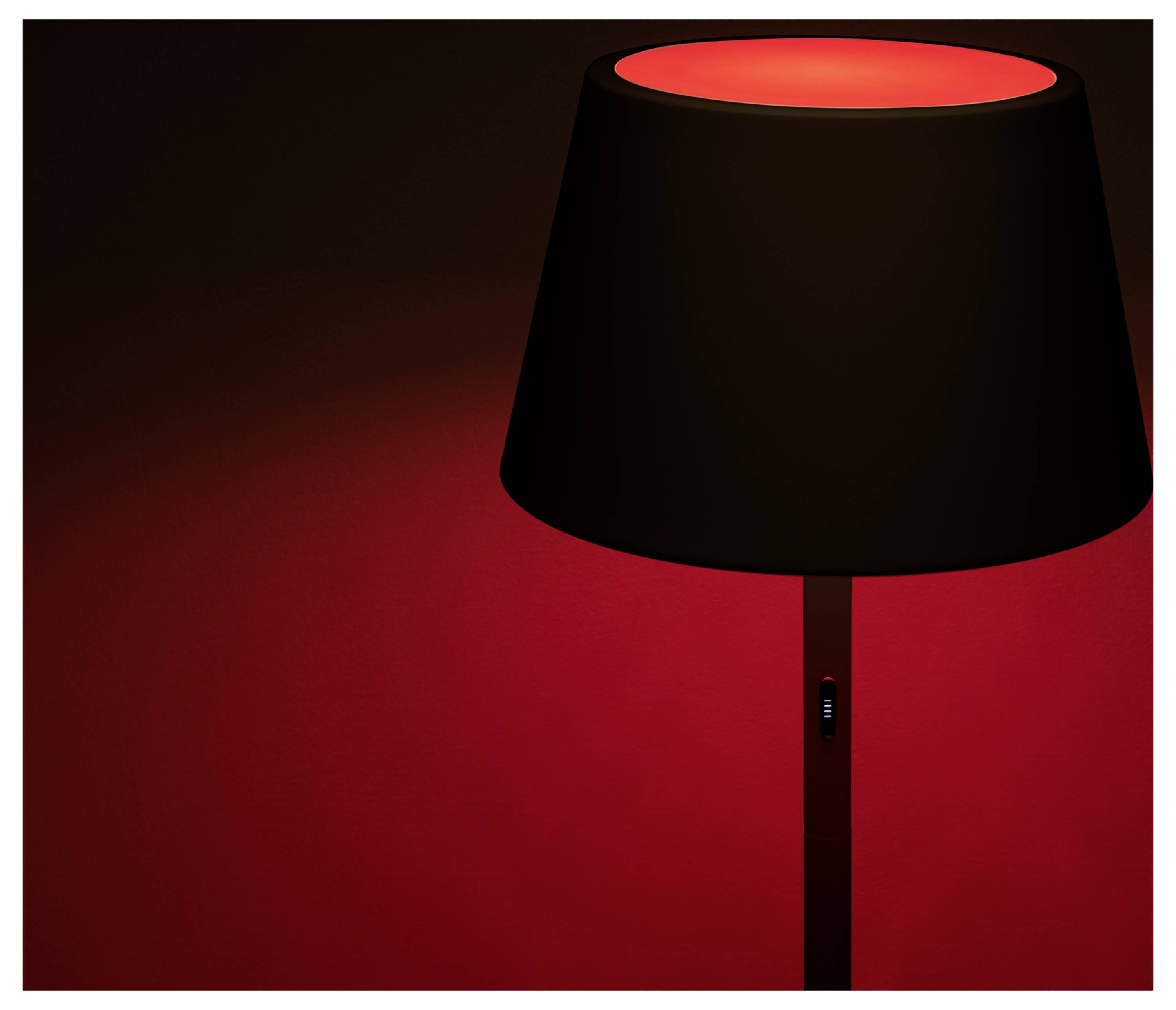 Eine Lampe mit einem schwarzen Lampenschirm wirft einen roten Schein vor einem dunklen Hintergrund und erzeugt eine warme und stimmungsvolle Atmosphäre.