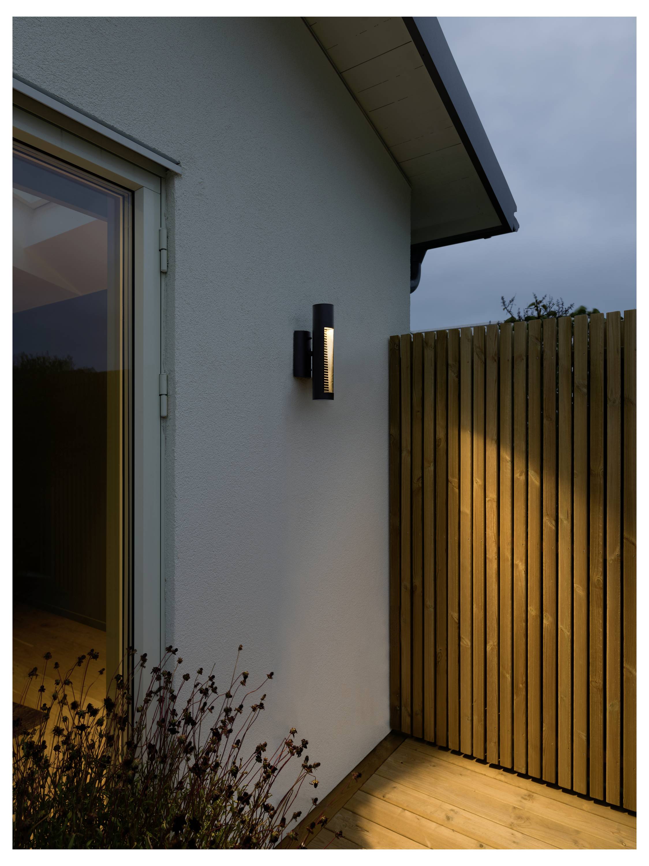 Eine moderne Wandleuchte ist außen an einem Holzzaun montiert und beleuchtet sanft einen kleinen Gartenbereich während der Abenddämmerung.