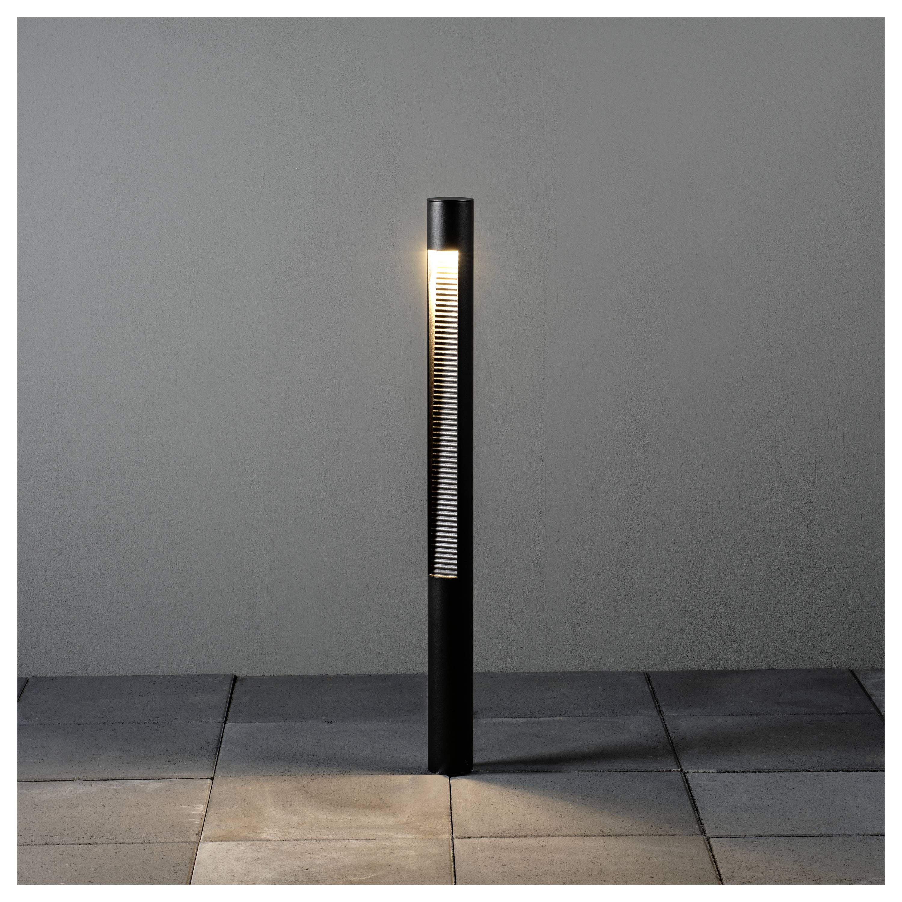 Eine schlichte, zylindrische Außenlampe auf einem Pfahl strahlt ein weiches, warmes Licht aus und steht auf einer gefliesten Terrasse vor einer einfarbigen grauen Wand.