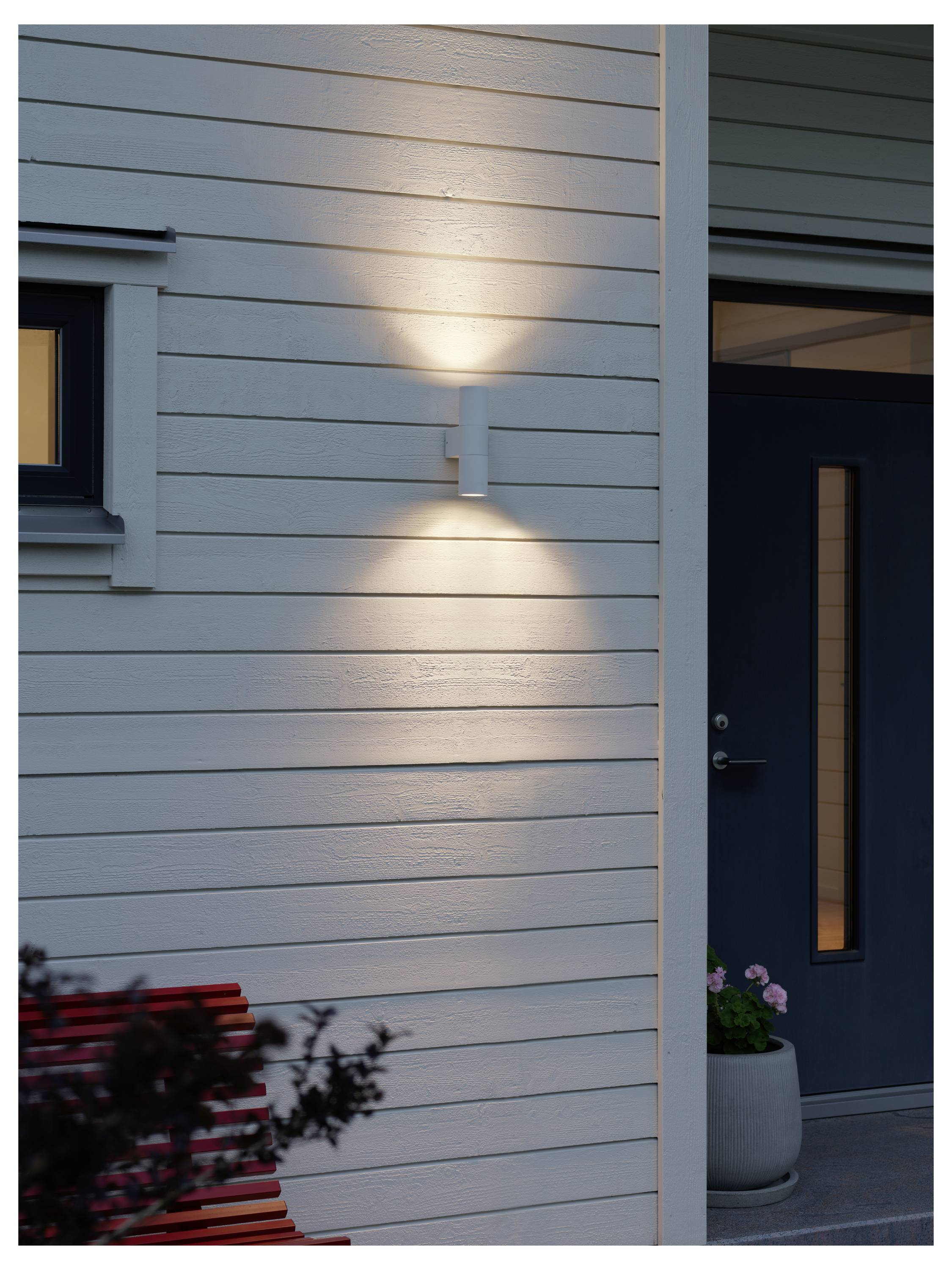 Moderne Außenwandleuchte, die bei Einbruch der Dämmerung die Außenfassade eines Hauses beleuchtet und an der horizontalen Holzverkleidung in der Nähe einer dunkelblau gestrichenen Tür mit einem Fenster montiert ist.