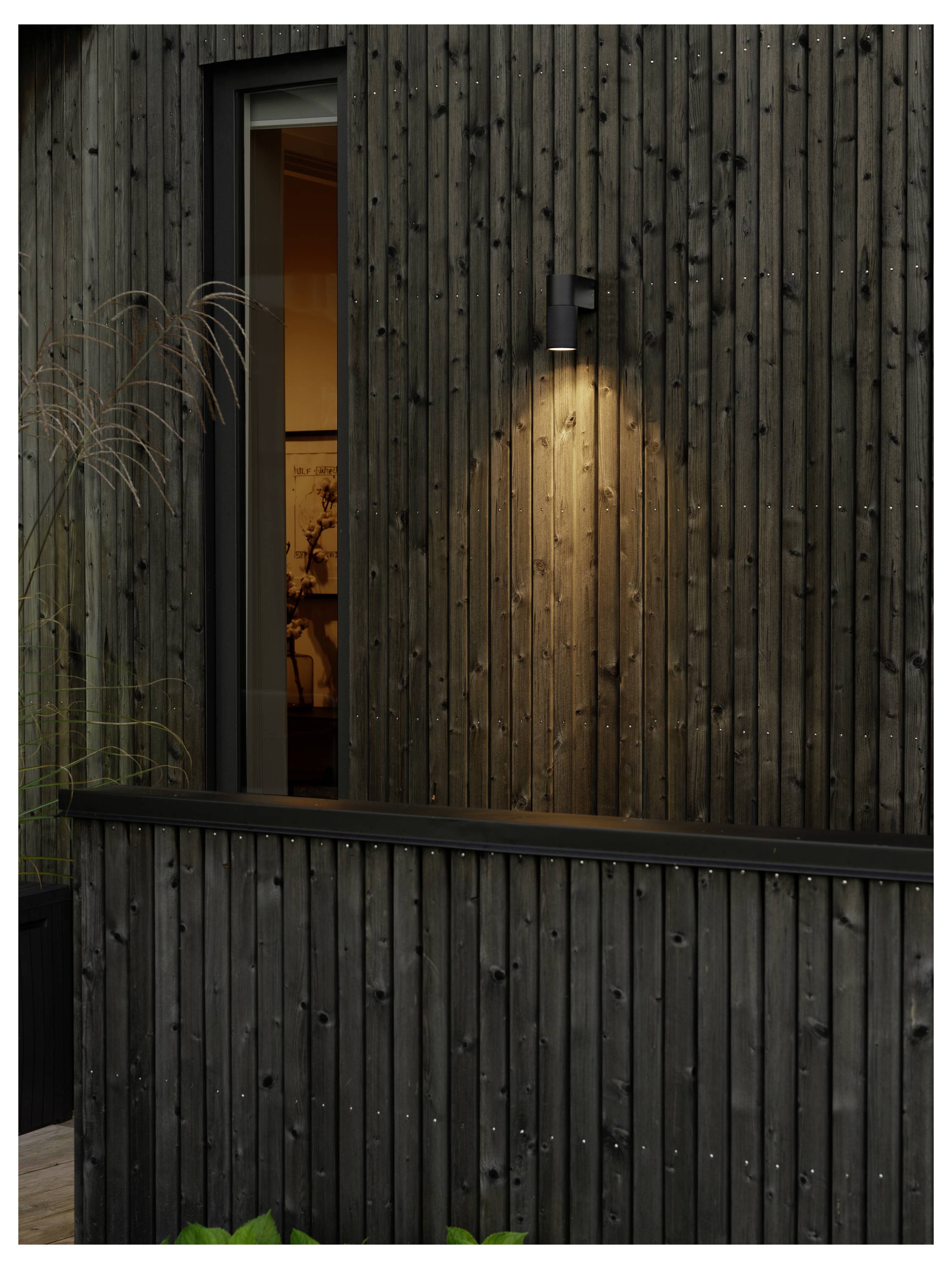 Eine Holzwand mit einer beleuchteten Außenlampe, die ein warmes Leuchten verbreitet. Ein Fenster gibt einen teilweise sichtbaren Innenraum mit blassen Dekorationen preis.