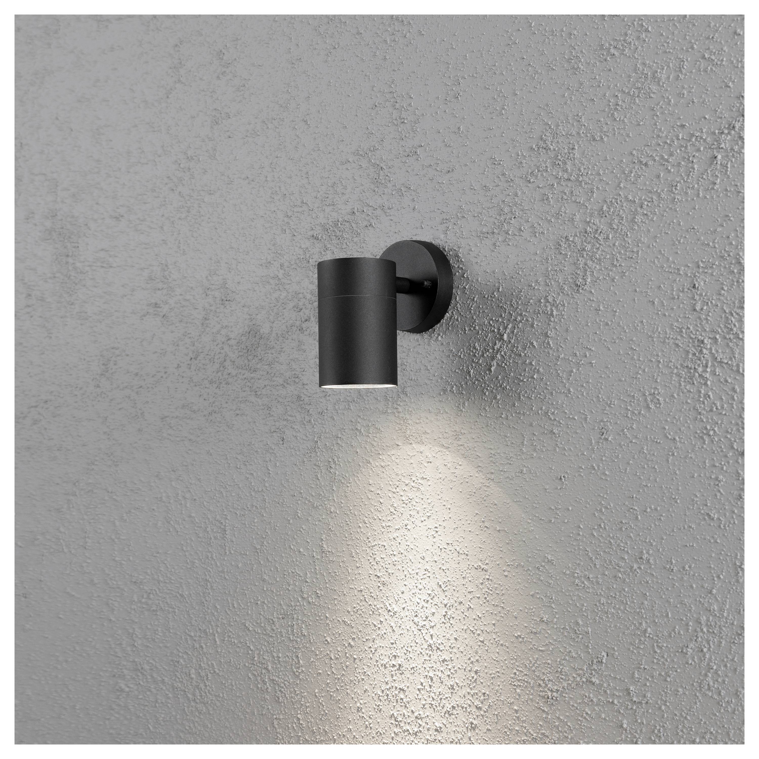 Eine schwarze, zylindrische Wandleuchte beleuchtet eine strukturierte graue Wand mit einem nach unten gerichteten Lichtstrahl.