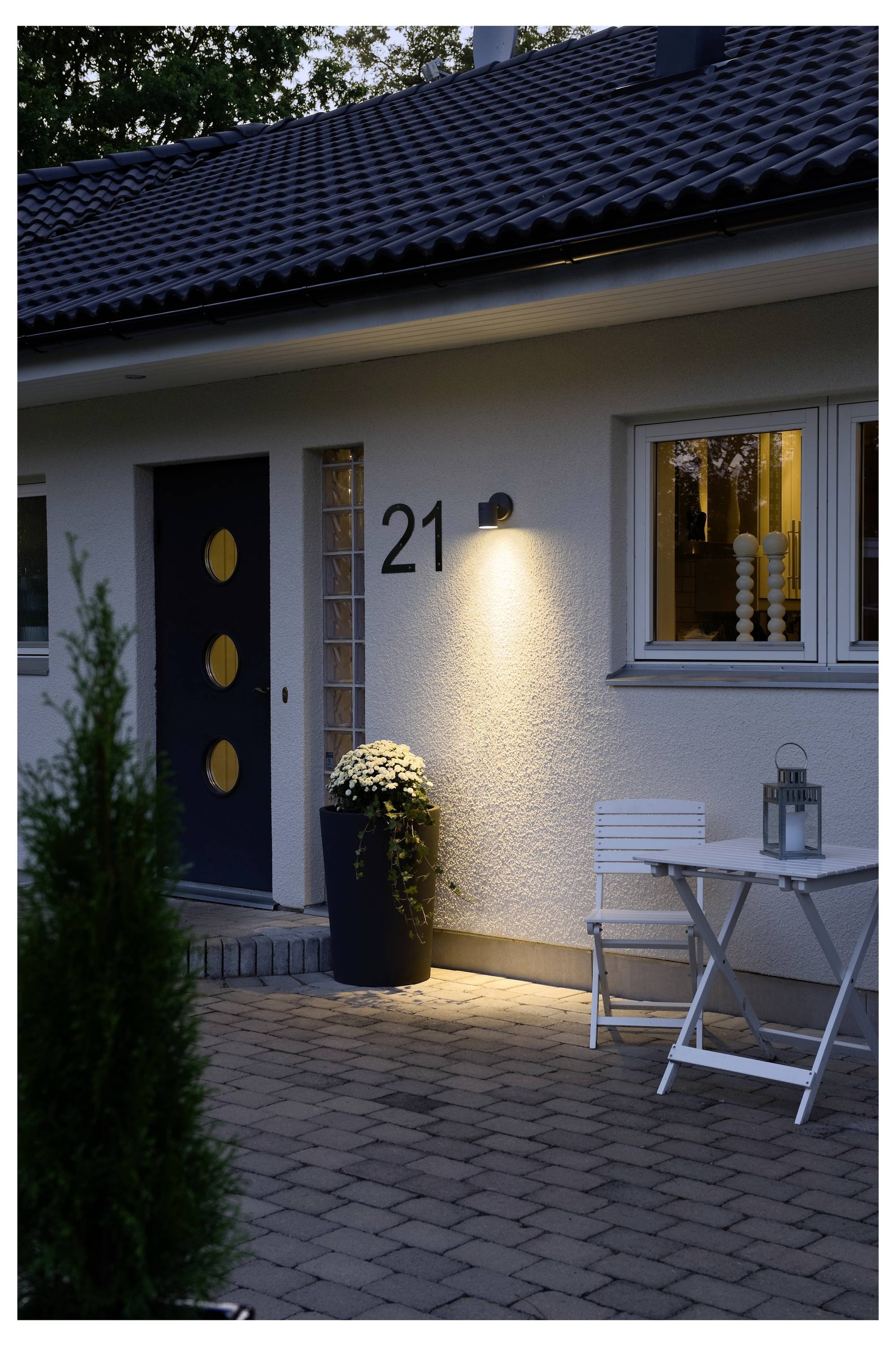 Eine moderne Hausfassade mit einer dunklen Tür, die Kreisfenster aufweist, der Hausnummer '21' an der Wand und einer kleinen Terrasse mit einem weißen Stuhl und Tisch.