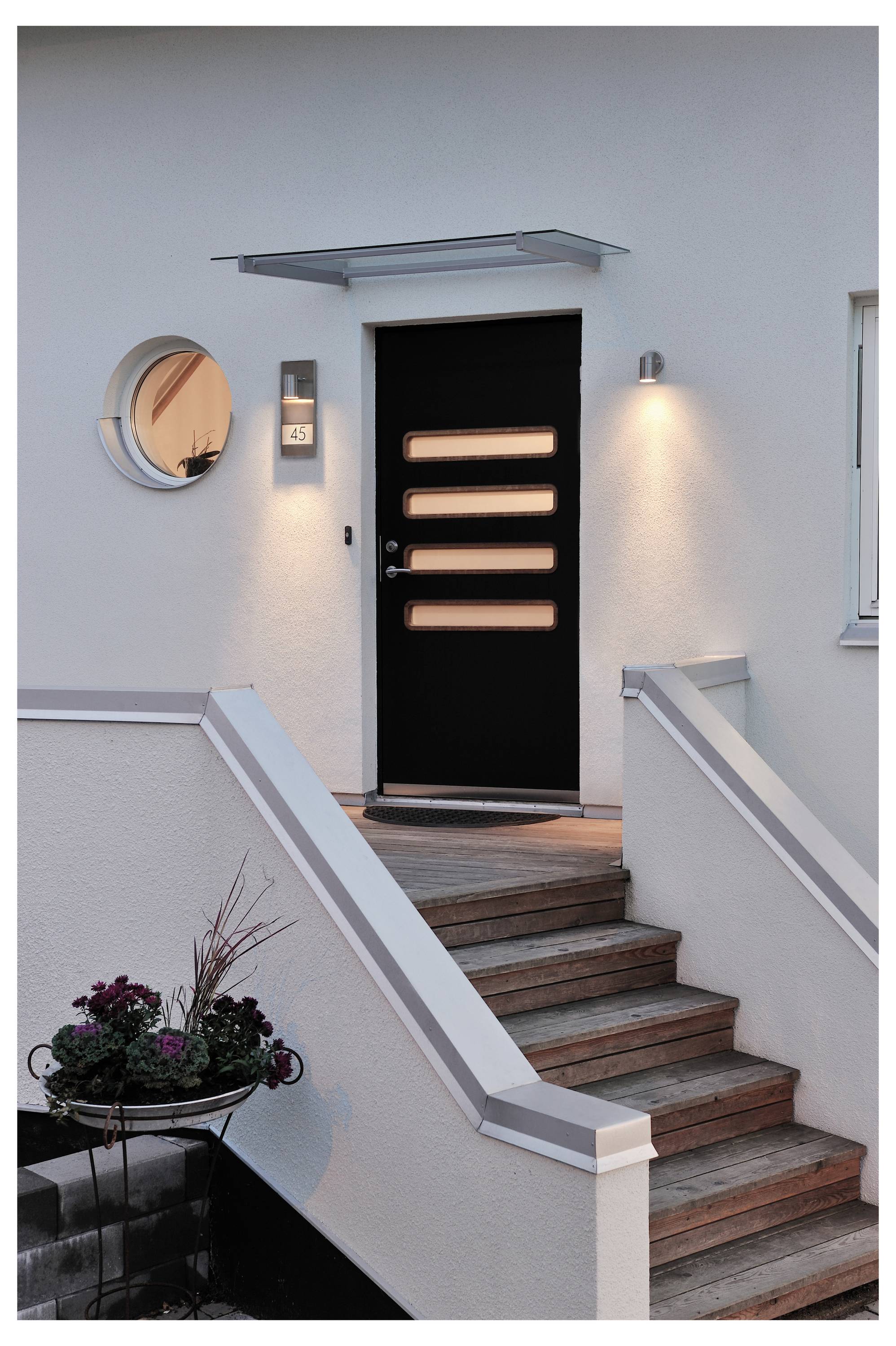 Moderner Hauseingang mit einer schwarzen Tür mit rechteckigen Fenstern, flankiert von beleuchteten Wandlampen und einer Treppe mit weißen Geländern, die nach oben führt.