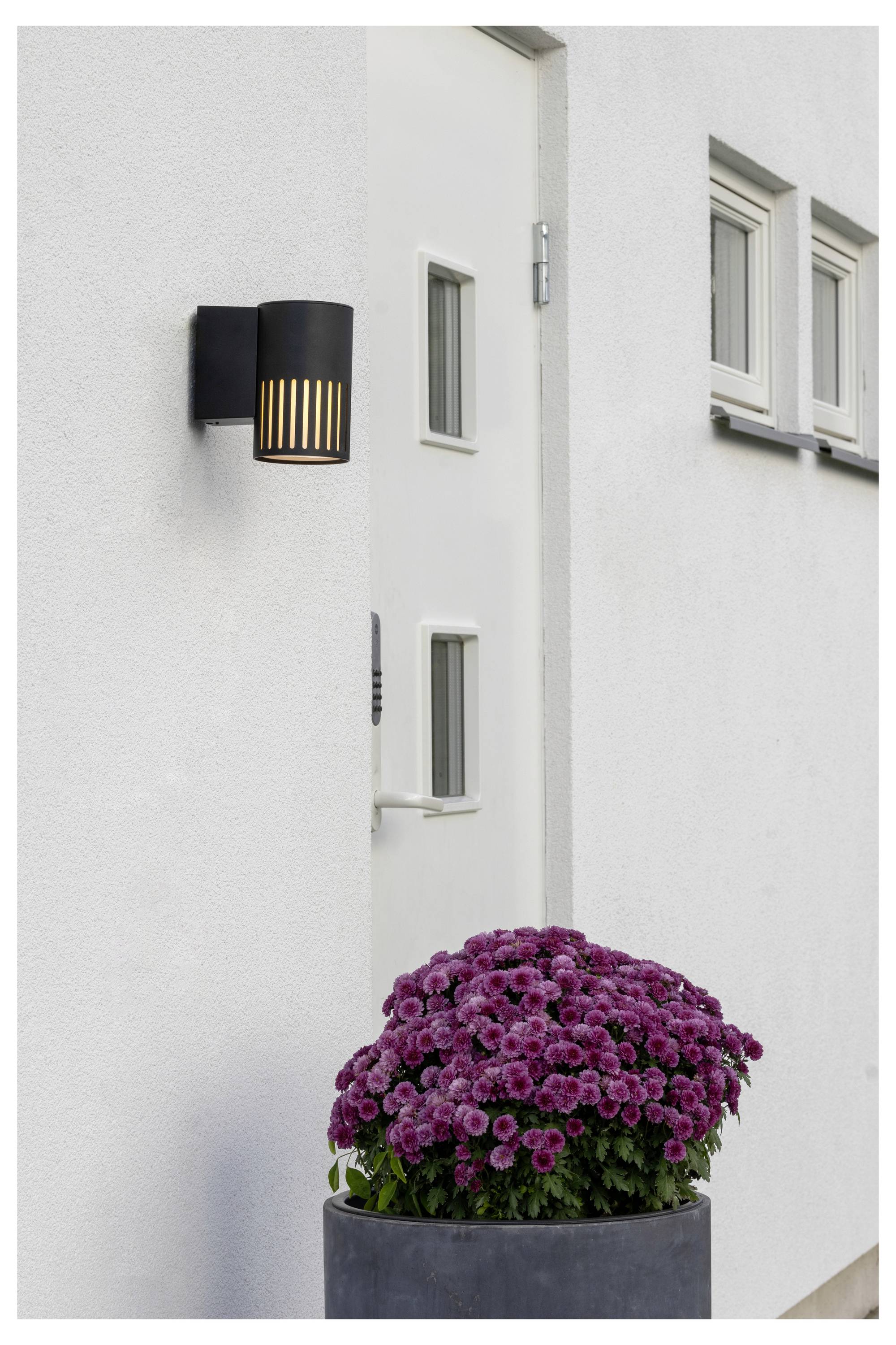 Eine moderne Außenleuchte neben einer weißen Tür, mit einem Topf voller lebhafter lila Blumen unterhalb von zwei kleinen Fenstern an einer weißen Wand.
