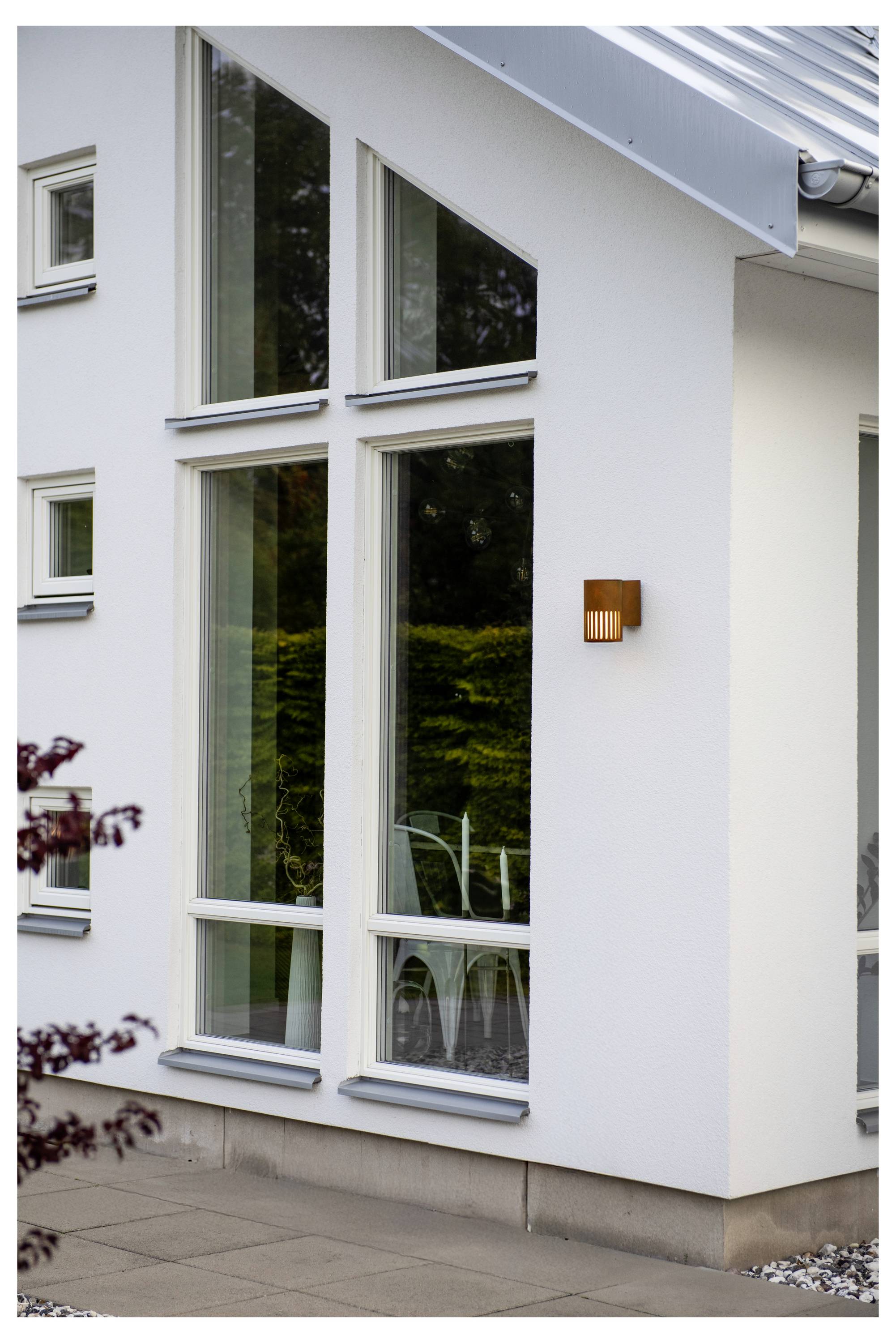 Modernes weißes Hausäußeres mit großen, vertikalen Fenstern, die Vegetation spiegeln; neben dem Eingang ist eine wandmontierte Leuchte sichtbar.