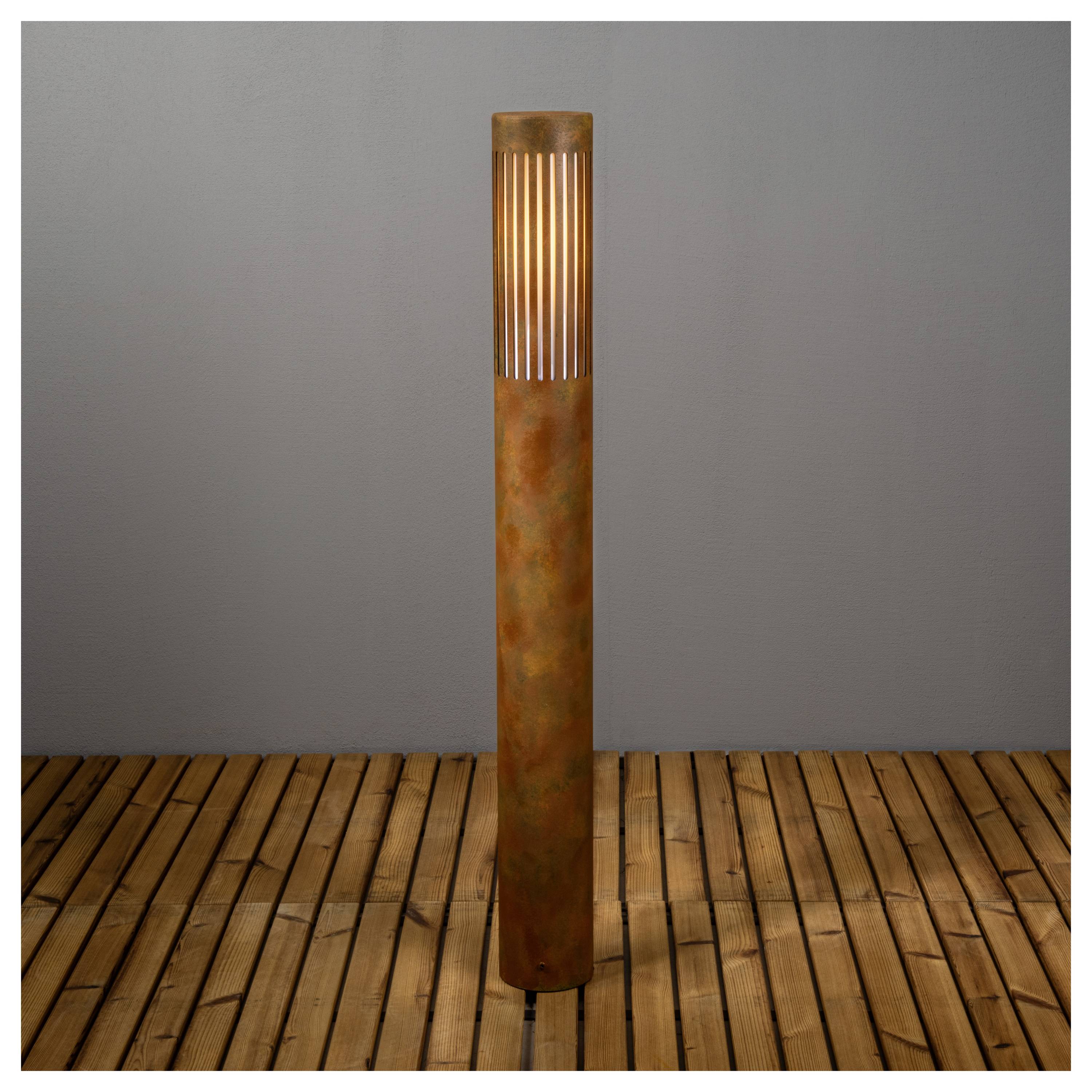 Eine hohe, rostfarbene Außenlampe mit einem leuchtenden Oberteil, die auf einer Holzterrasse vor einer schlichten Wand steht.