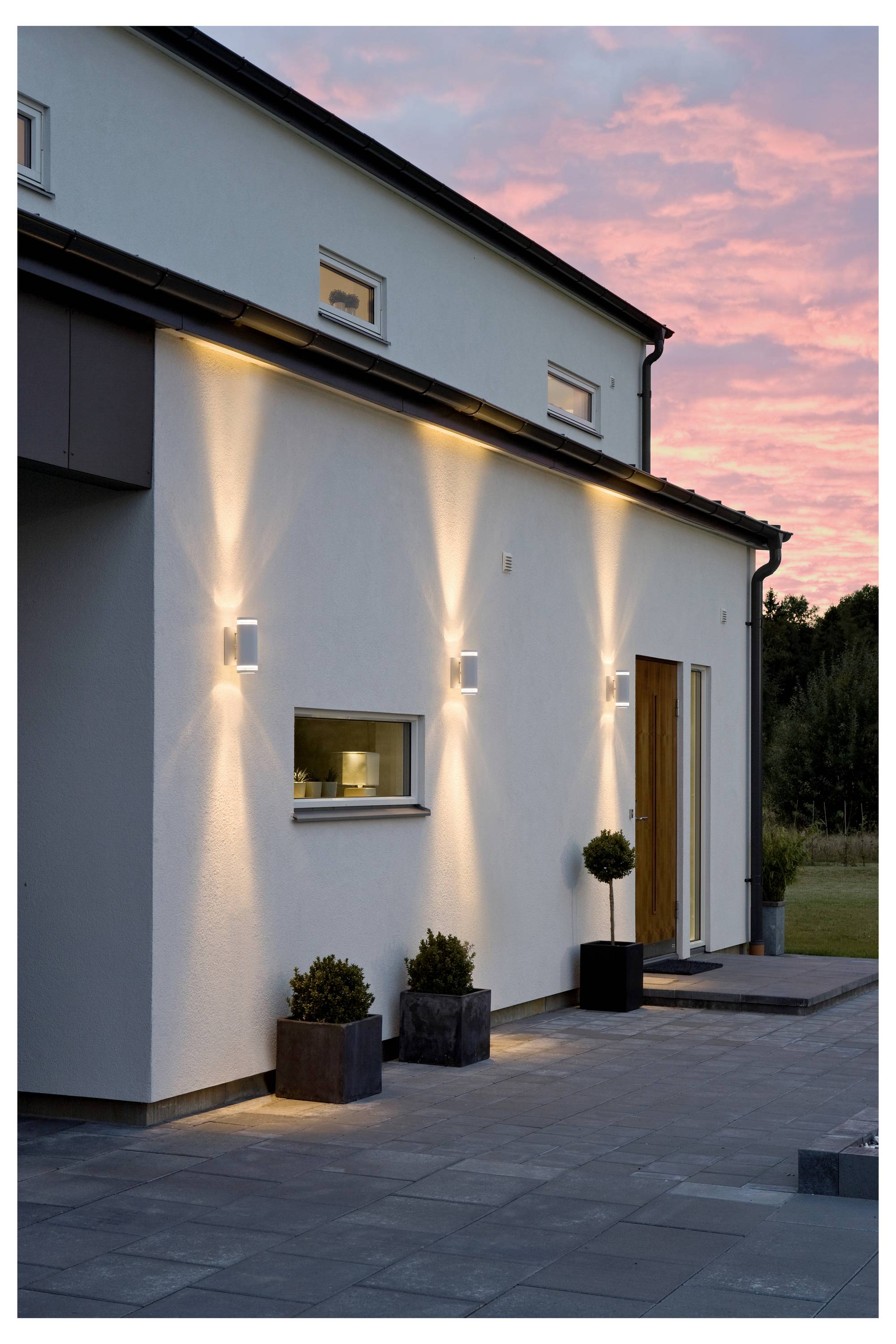 Zeitgenössisches Hausäußeres bei Dämmerung mit beleuchteten Wandleuchten, moderner Architektur, großem Fenster und Topfpflanzen nahe dem Eingang.