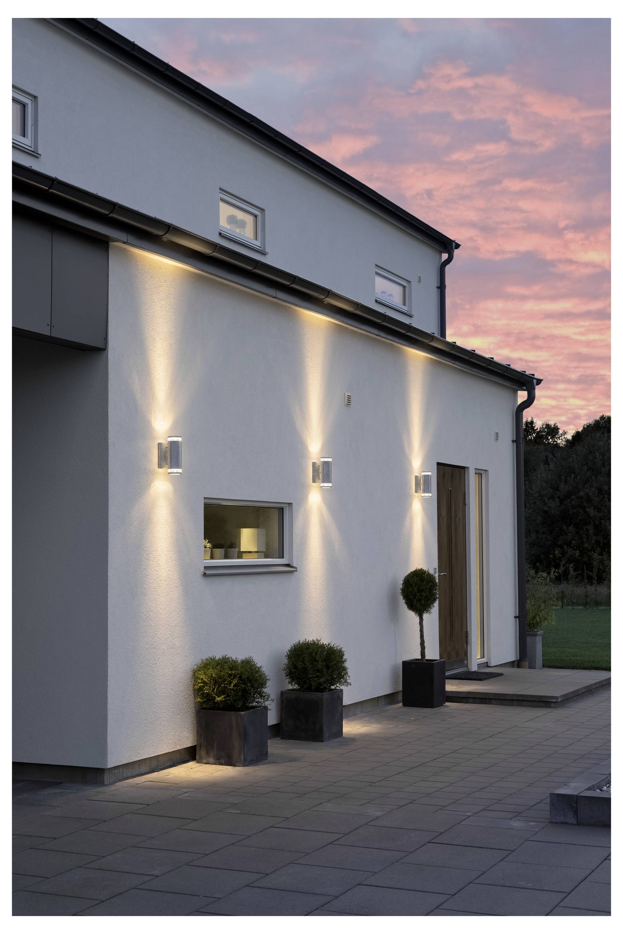 Moderne Hausfassade mit drei vertikalen Wandleuchten, kleinen Pflanzen in Töpfen in der Nähe des Eingangs und einem Abendhimmel im Hintergrund.