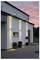 Moderne Hausfassade mit drei vertikalen Wandleuchten, kleinen Pflanzen in Töpfen in der Nähe des Eingangs und einem Abendhimmel im Hintergrund.