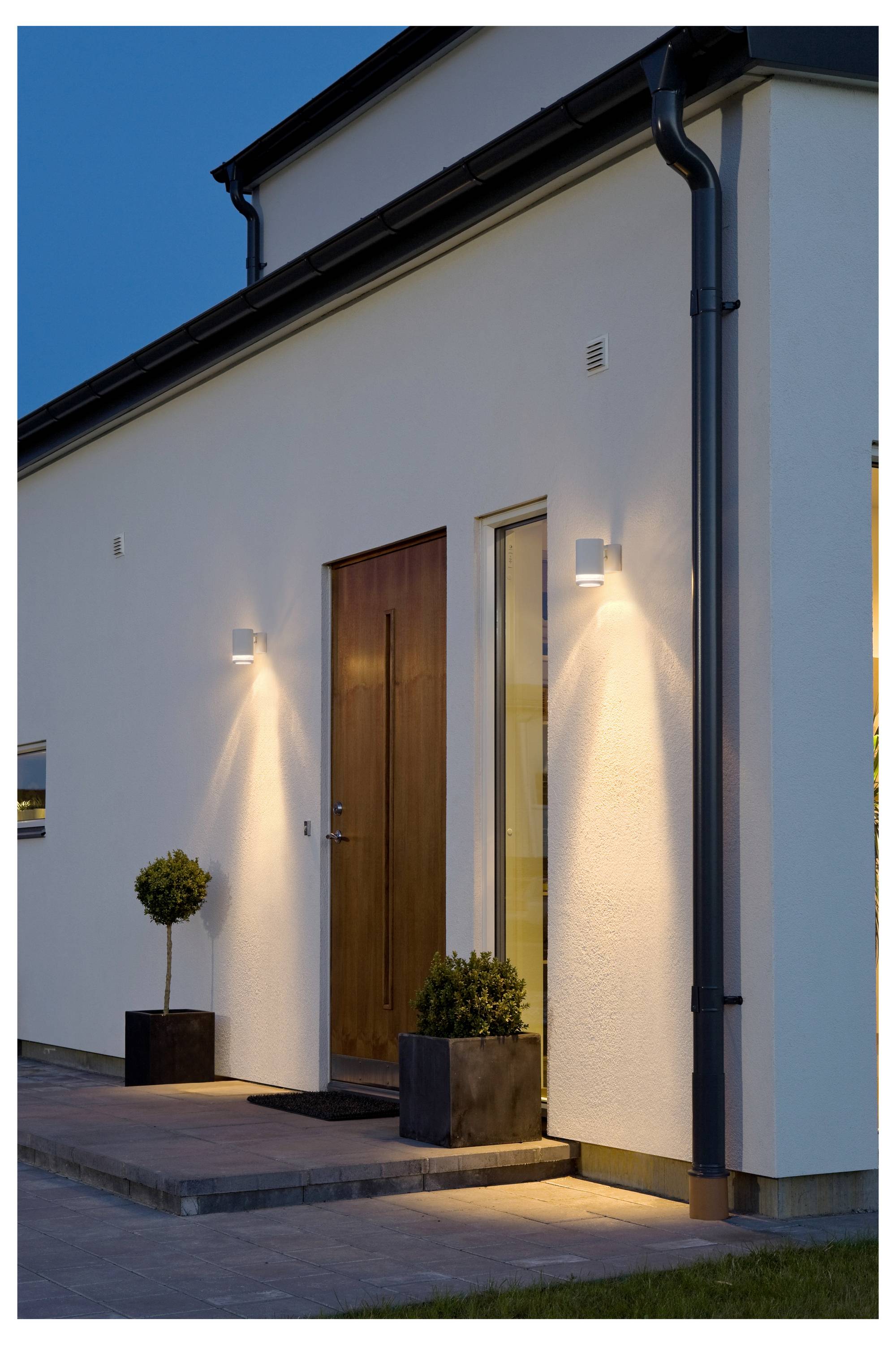 Eine moderne Hauseinfahrt in der Dämmerung mit einer Holztür, zwei kleinen Kübelpflanzen und wandmontierter Beleuchtung, die die Fassade erleuchtet.